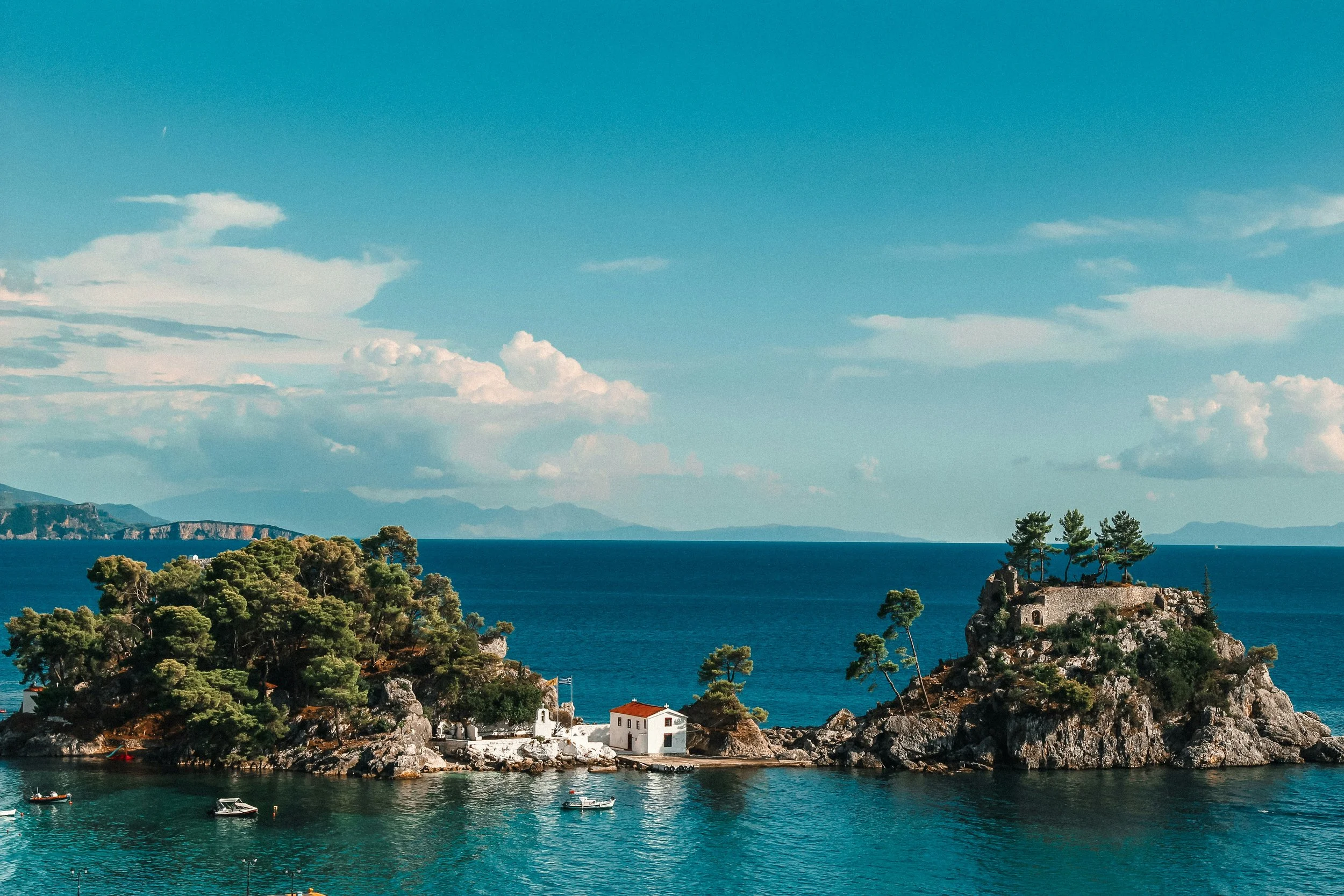 A scenic coastal landscape with a calm turquoise sea, rocky shoreline with trees, and a small white building with a red roof, possibly a church or house, near the water's edge. In the background are distant mountains and a partly cloudy sky.