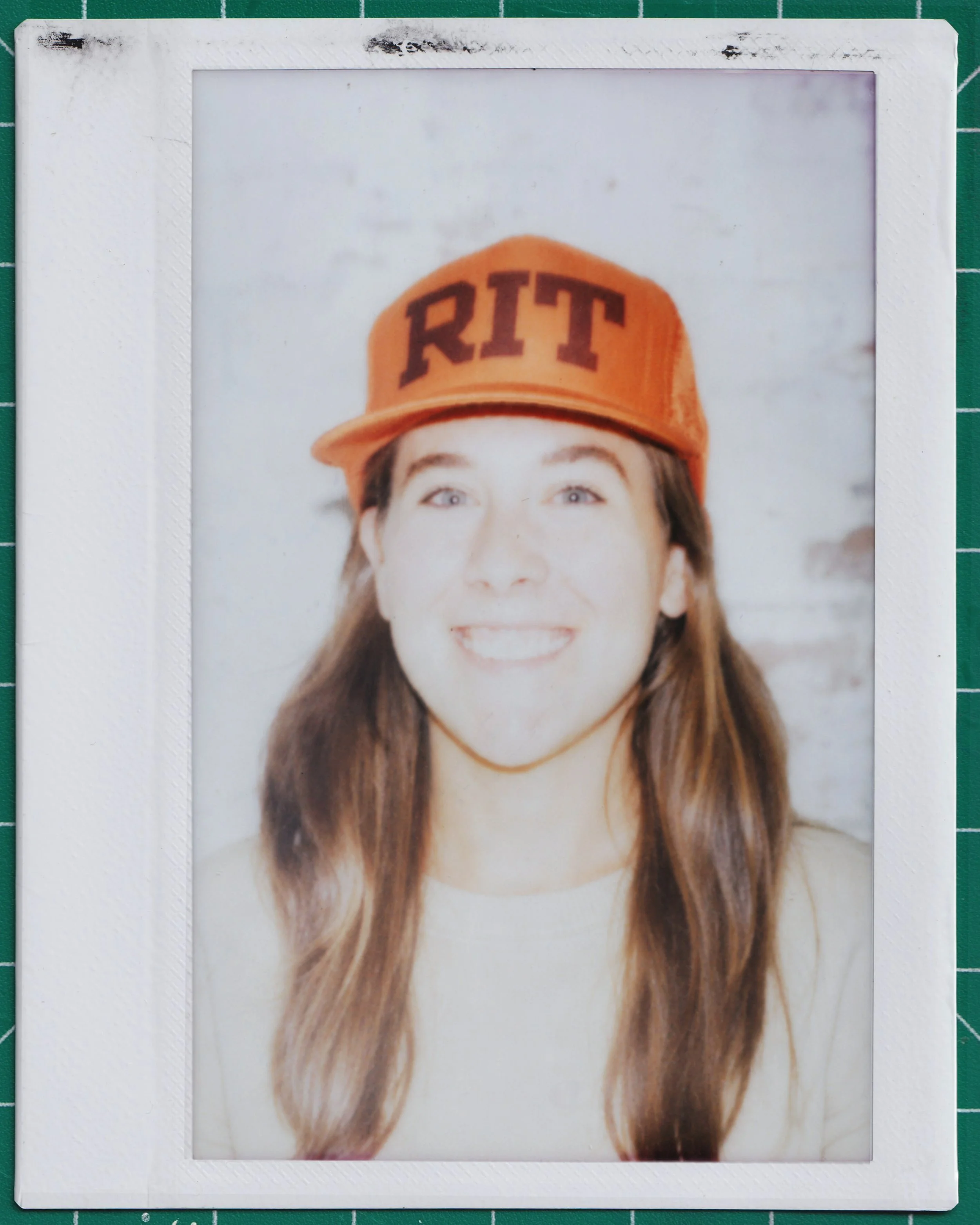 Polaroid portrait of Alyssa Barnes in an orange RIT cap, photographed on a green cutting mat.