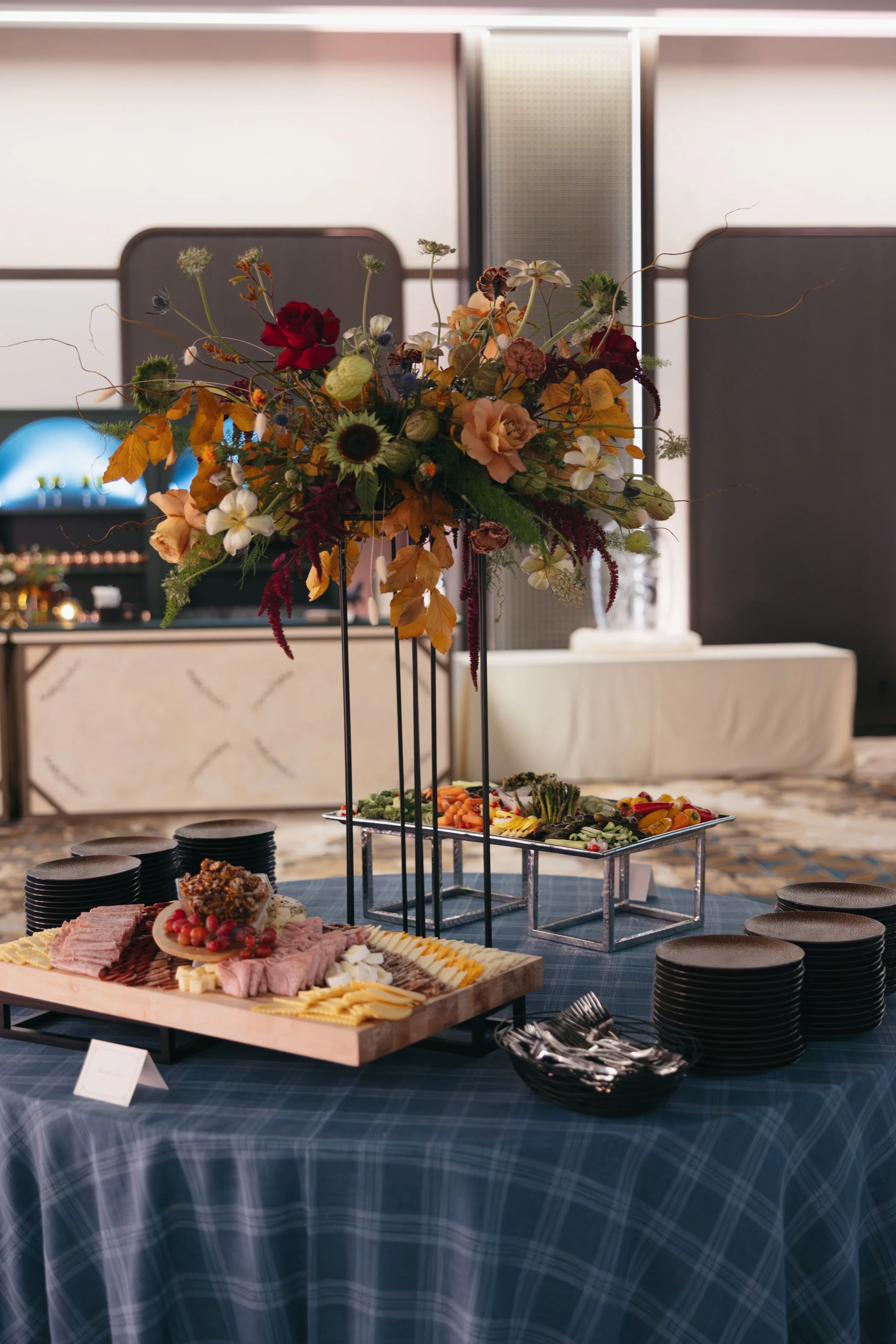 Buffet table with a large floral centerpiece, featuring orange, red, white, and purple flowers, and a variety of cheeses, meats, and vegetables arranged on platters, set on a blue checkered tablecloth.