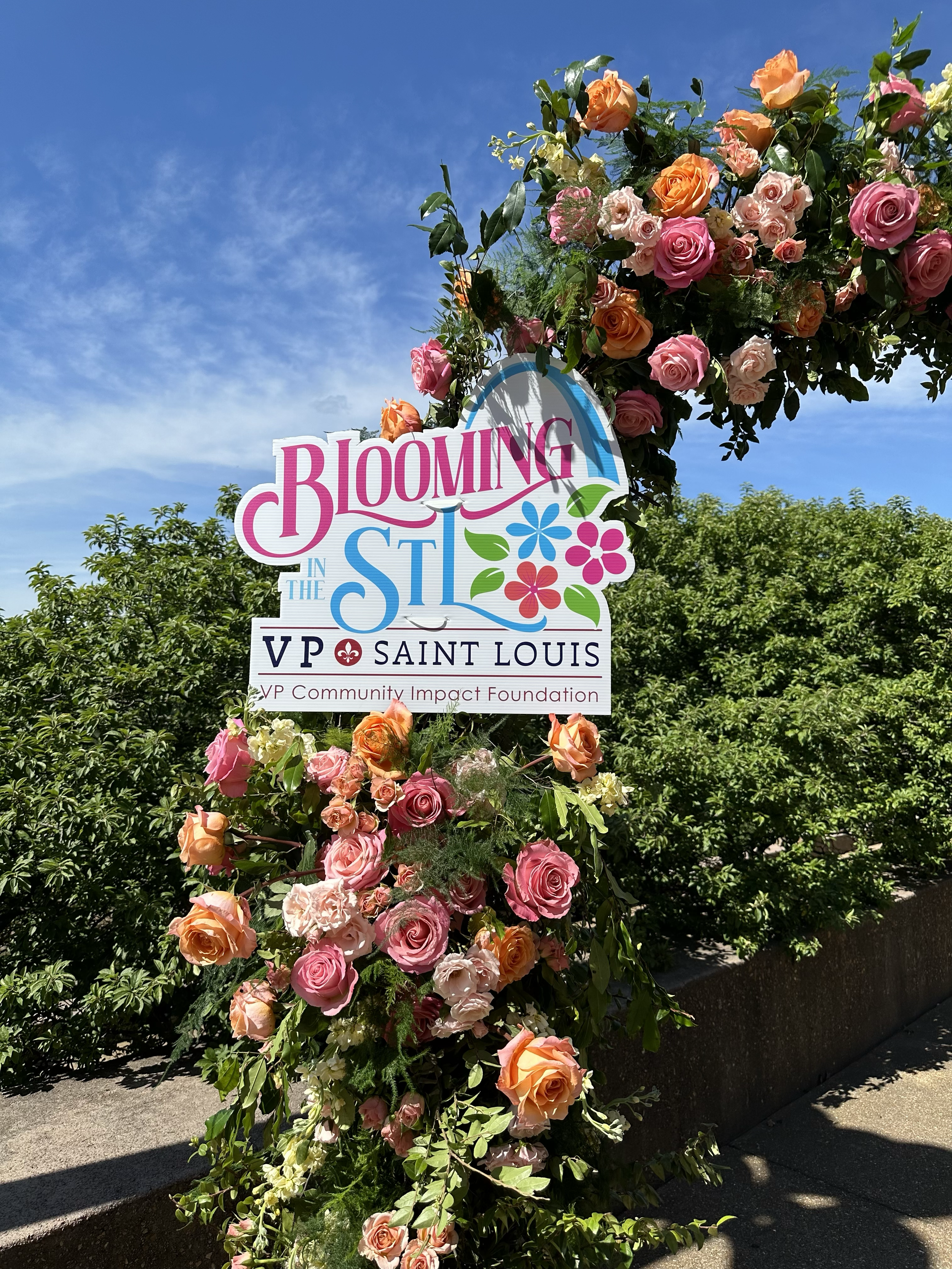 A floral arrangement with pink, orange, and cream roses and greenery outdoors under a blue sky with some clouds. The arrangement includes a sign for 'Blooming in the St Louis VP Saint Louis' and 'VP Community Impact Foundation' at the base.