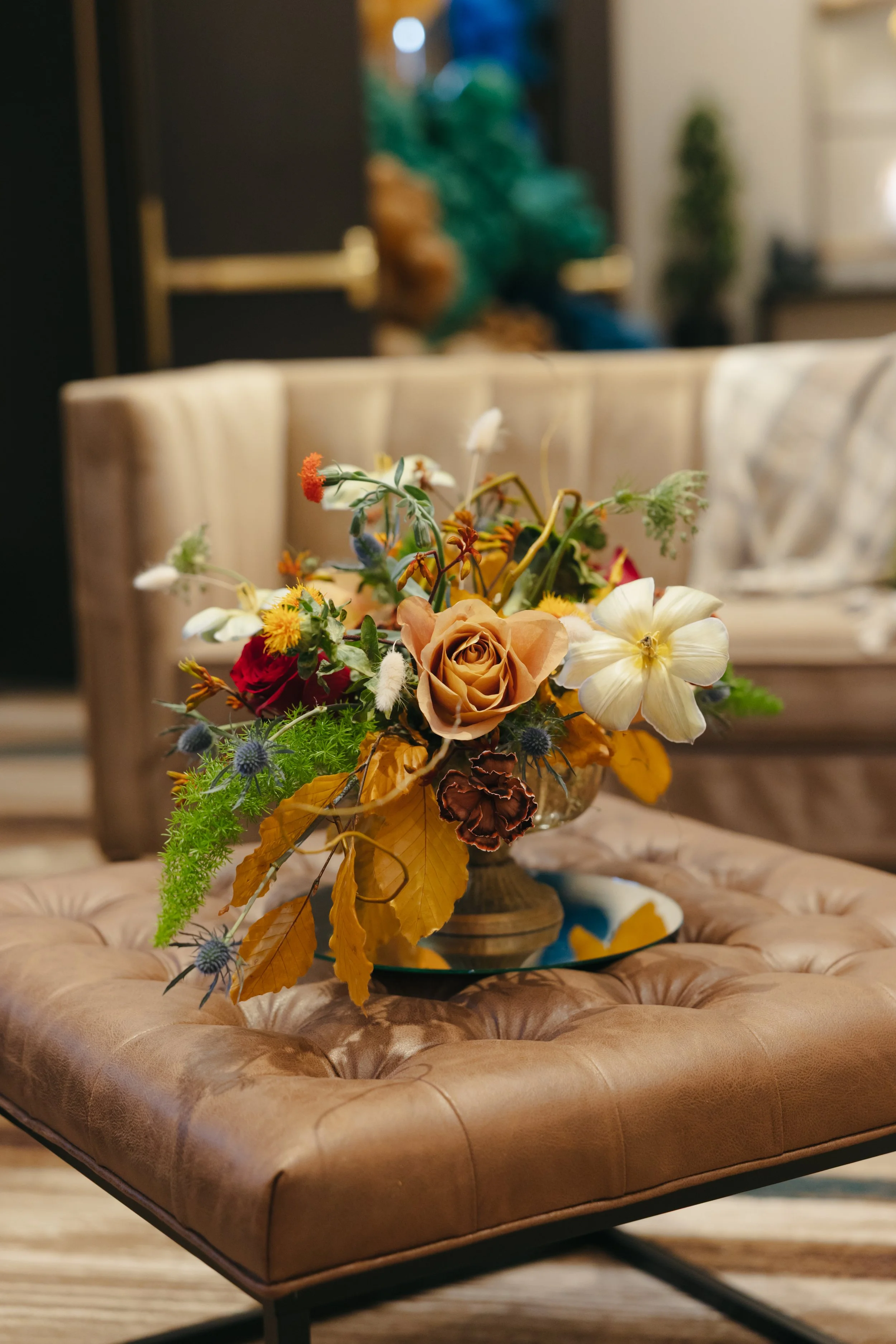 A bouquet of artificial flowers with beige, white, yellow, red, green, and blue colors placed on a tufted tan leather ottoman in a warmly lit room.