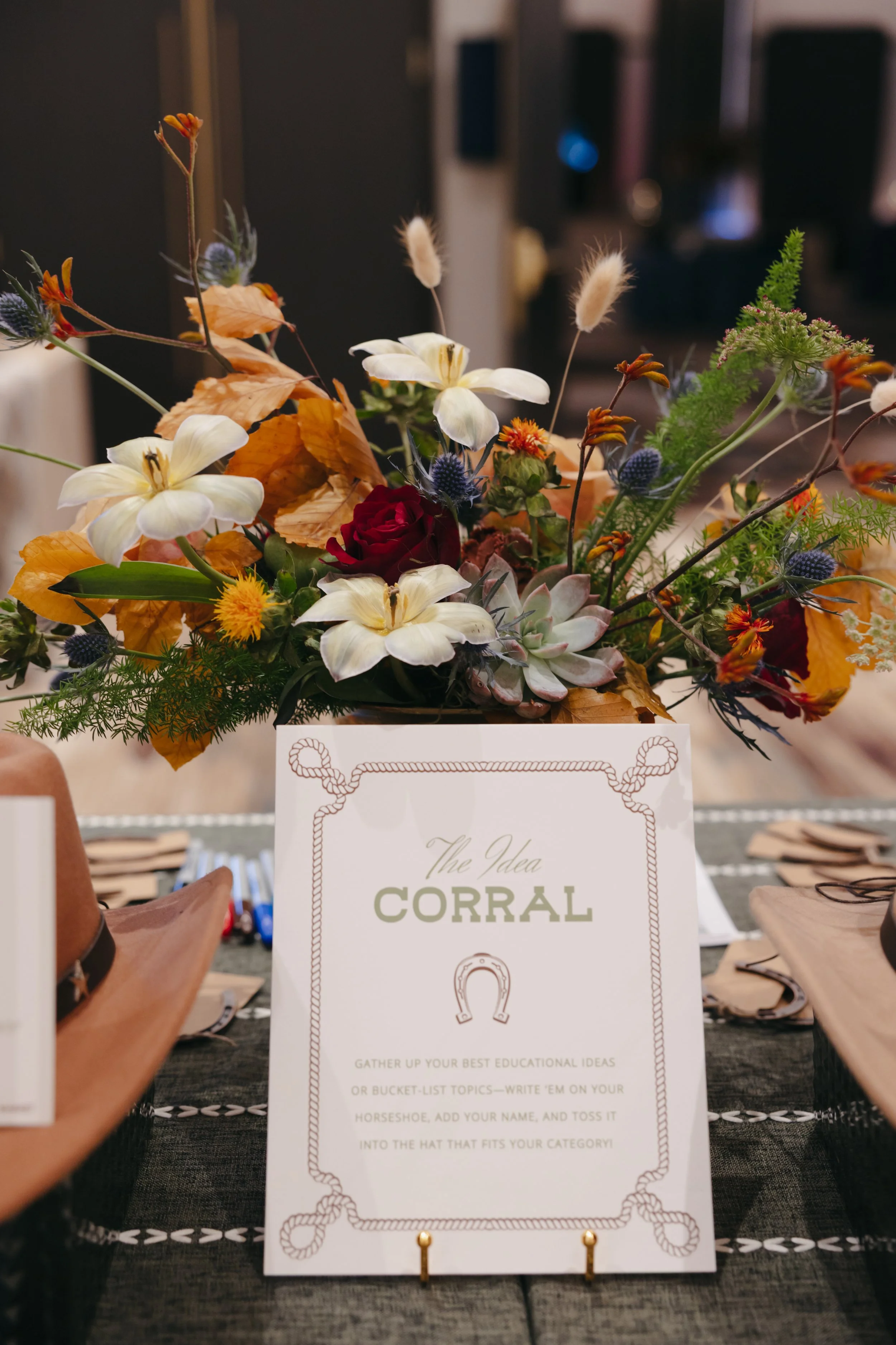A floral arrangement with white lilies, red roses, yellow and orange flowers, dried leaves, and green foliage on a table. In front of the arrangement, there is a white card with green text labeled 'The Idea' from 'Corral,' which includes instructions