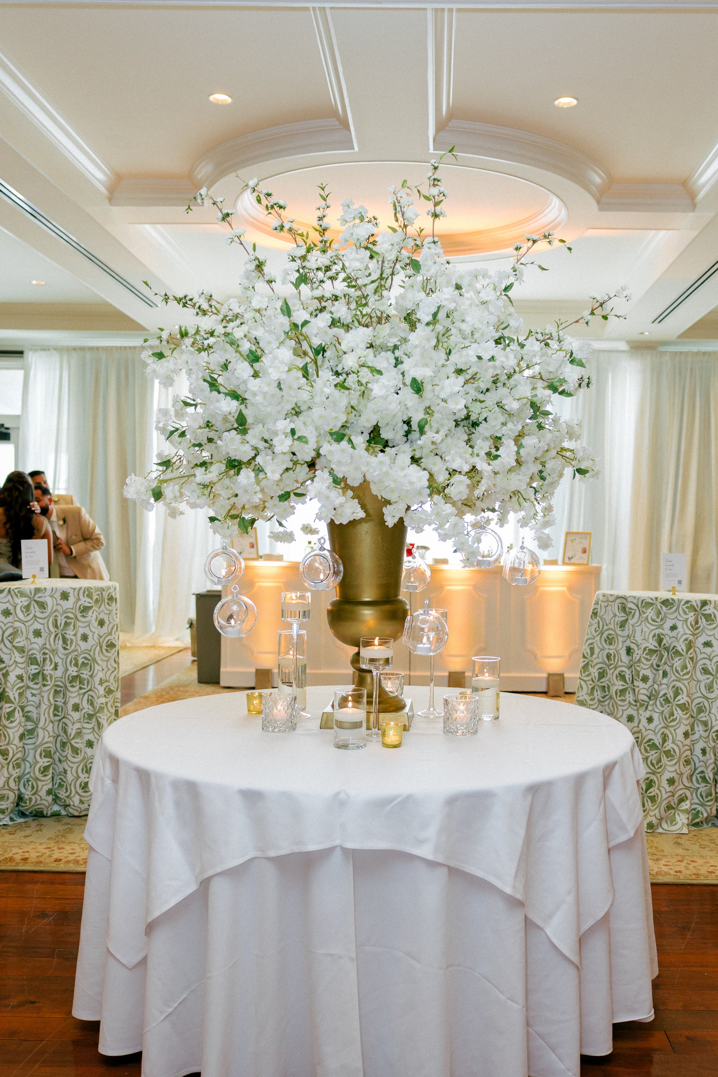Round table with a white tablecloth holding a large gold vase filled with white flowers, surrounded by candles, in a decorated banquet hall.