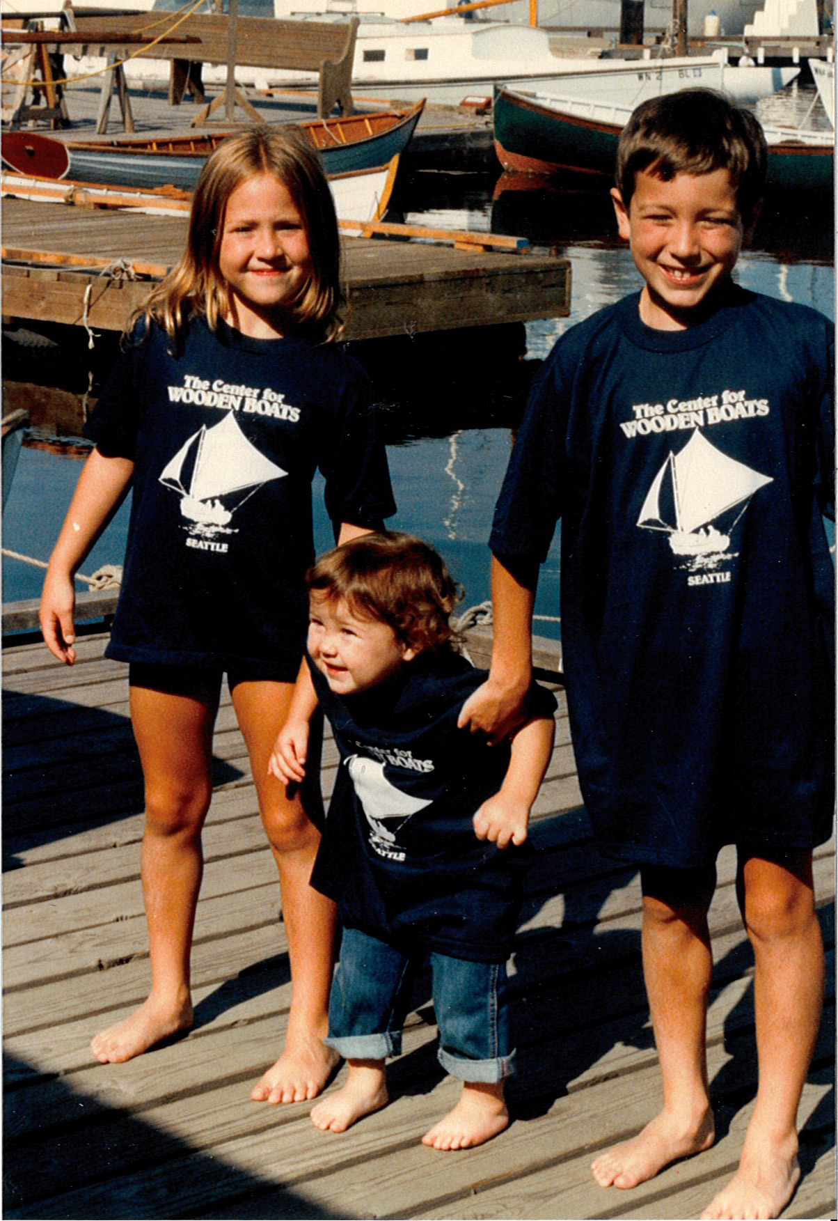 Three smiling children are standing on the dock on a sunny day