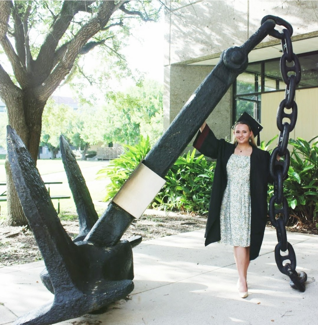 Libby Barnes is wearing a graduation cap and gown, is standing by a large anchor sculpture