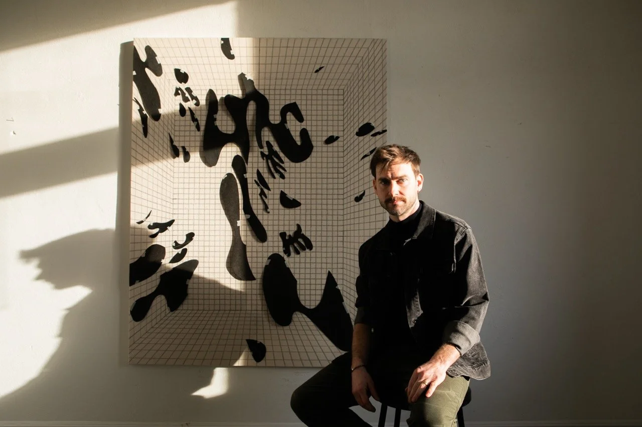 A man with a beard and mustache sits on a stool in front of an abstract art piece with black shapes on a grid-patterned background, illuminated by sunlight and shadows.
