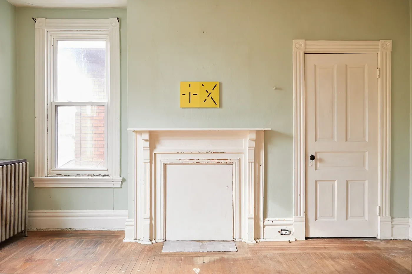 Interior of a room with a window on the left, a fireplace in the center, and a door on the right. The room has light green walls, wooden floors, and white trim.