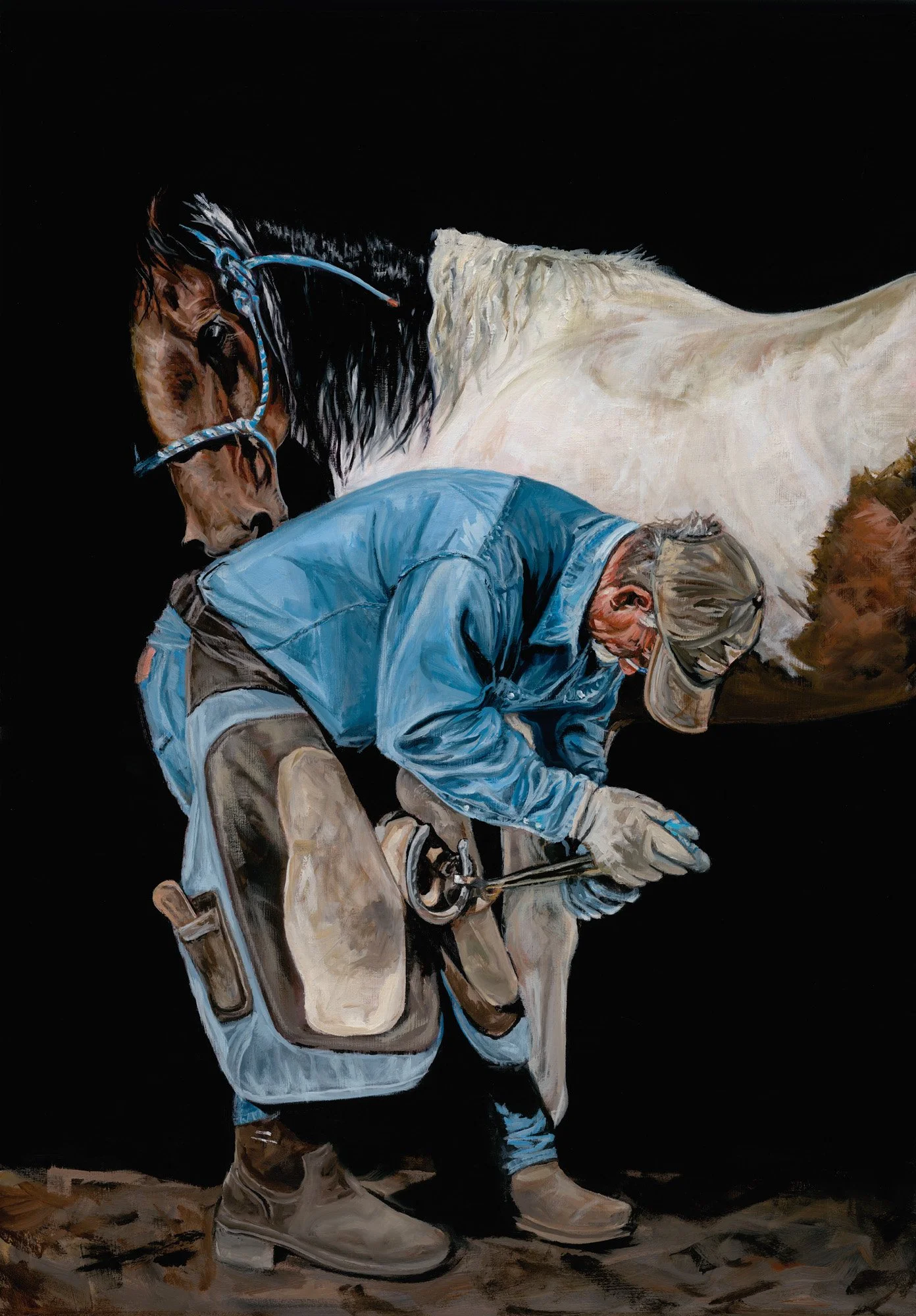 Farrier in blue jeans shoeing a pinto horse with dramatic lighting
