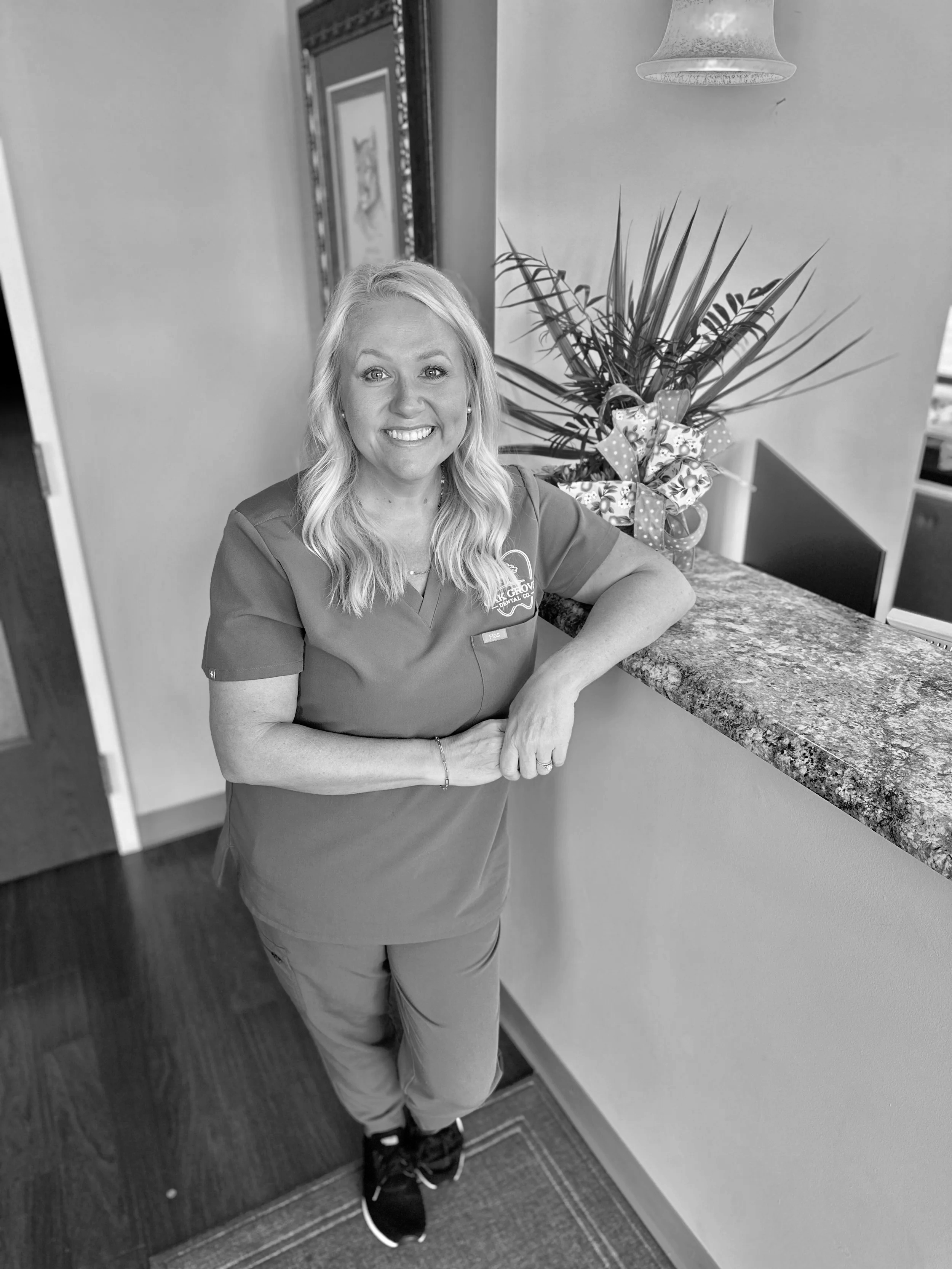 A woman with long wavy blonde hair smiling while standing at a reception desk, wearing medical scrubs, with a floral gift bag and a potted plant in the background.
