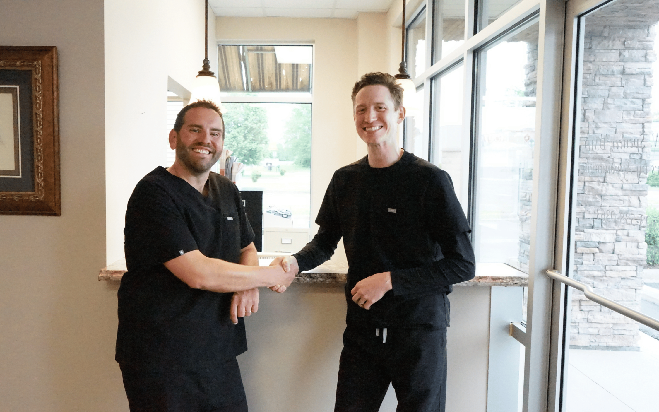 Two men in black scrubs shaking hands inside a building near a window with stone wall outside.
