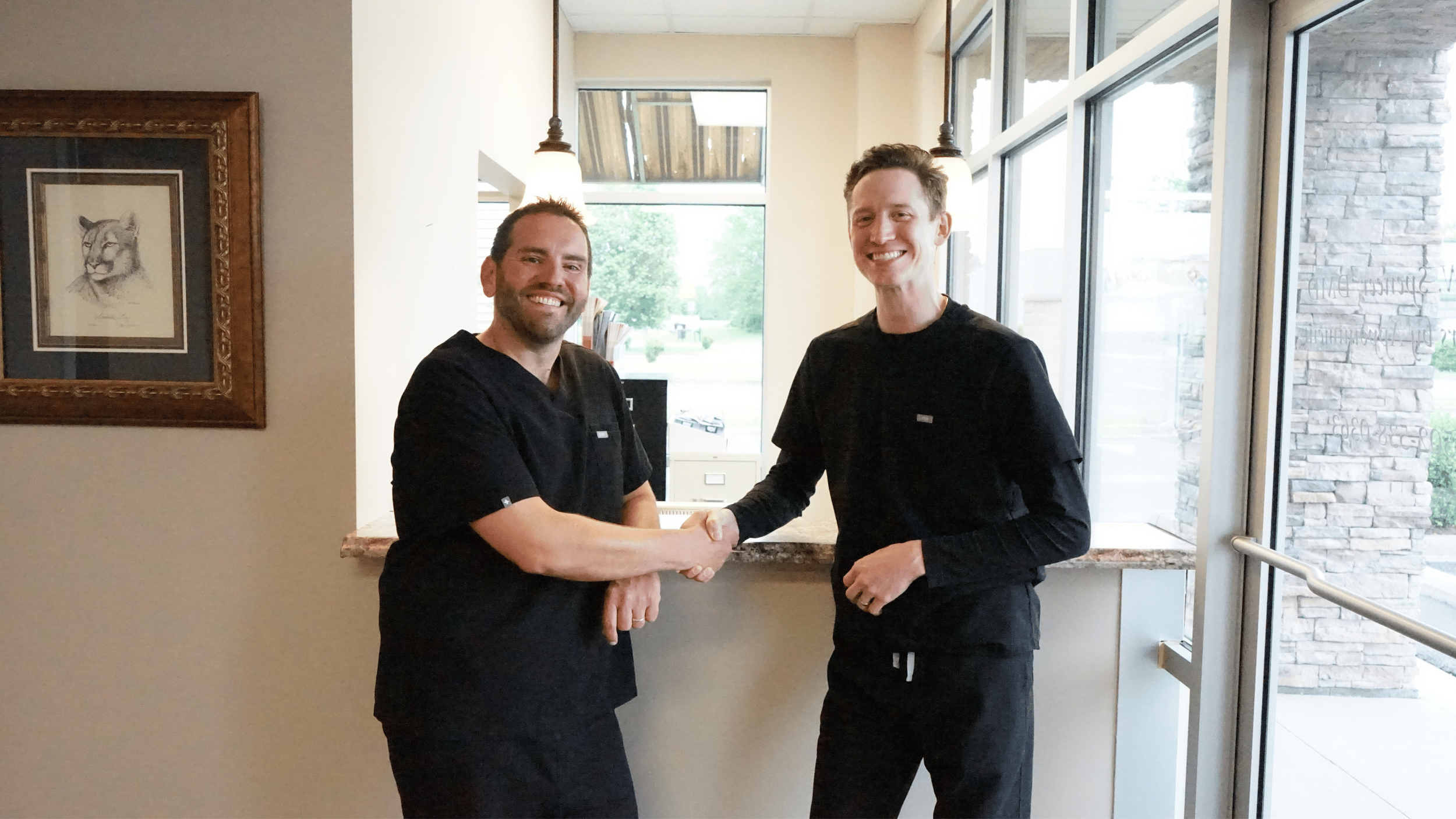 Two men with short hair and beards, wearing black scrubs, shaking hands and smiling inside a well-lit building with large glass windows.