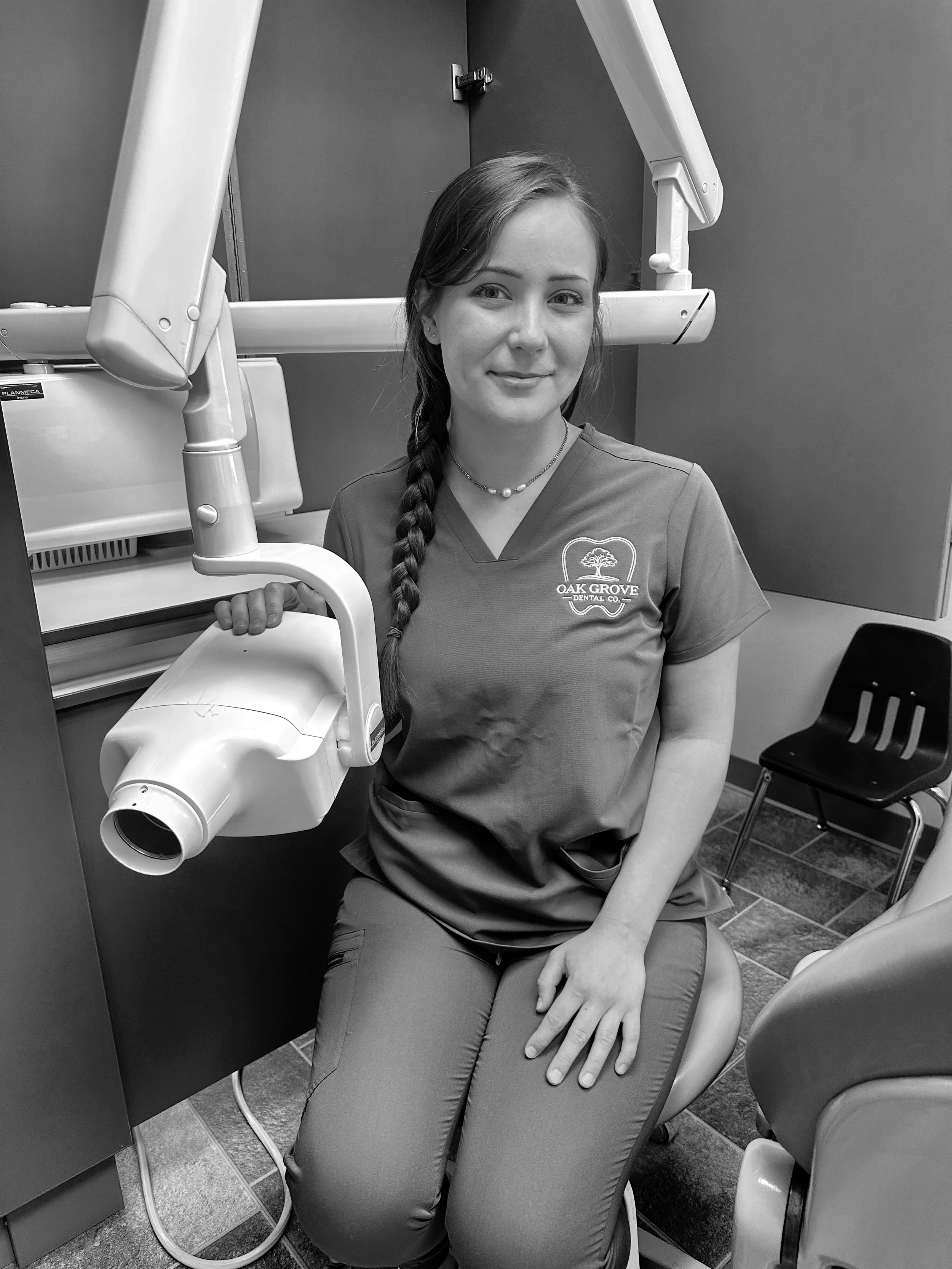 A woman in dental scrubs sitting in a dentist's office, with dental equipment around her and a chair in the background.