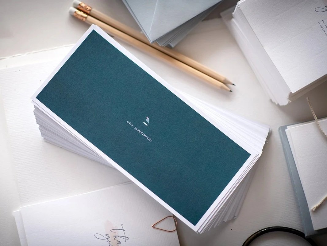 A stack of greeting cards with a card in front that has a teal cover and the words "with compliments" printed on it, surrounded by papers, pencils, and office supplies on a white desk.