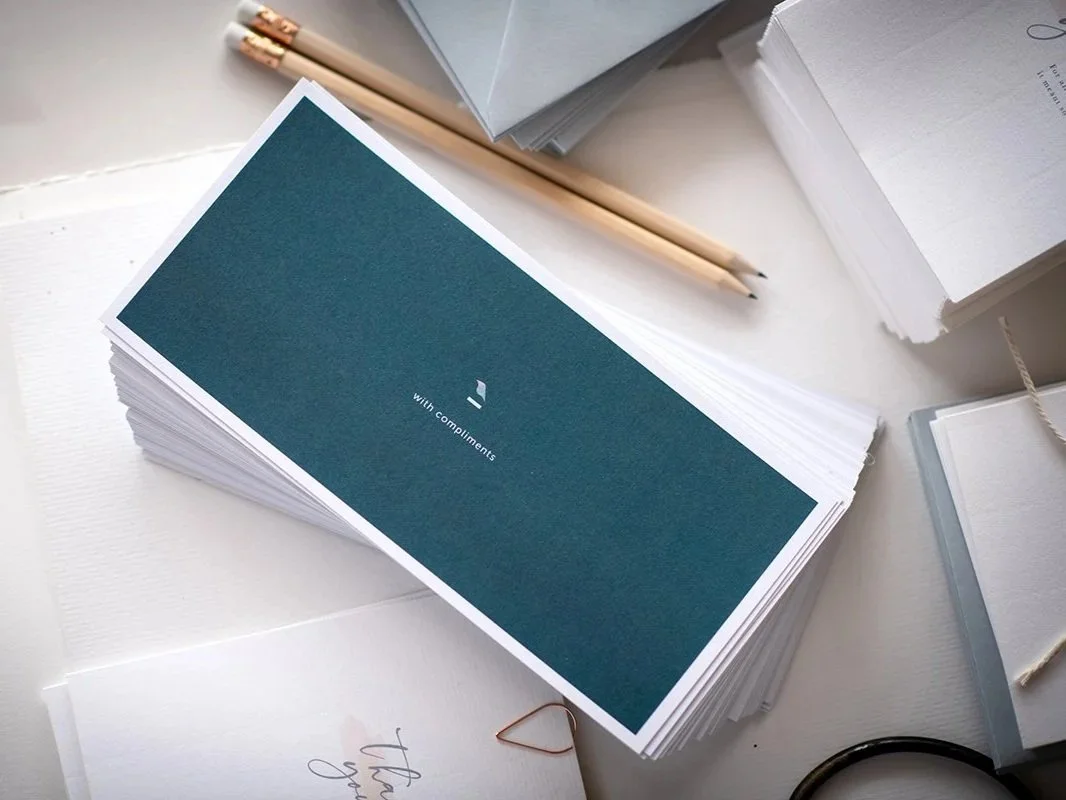 A stack of greeting cards with a card in front that has a teal cover and the words "with compliments" printed on it, surrounded by papers, pencils, and office supplies on a white desk.