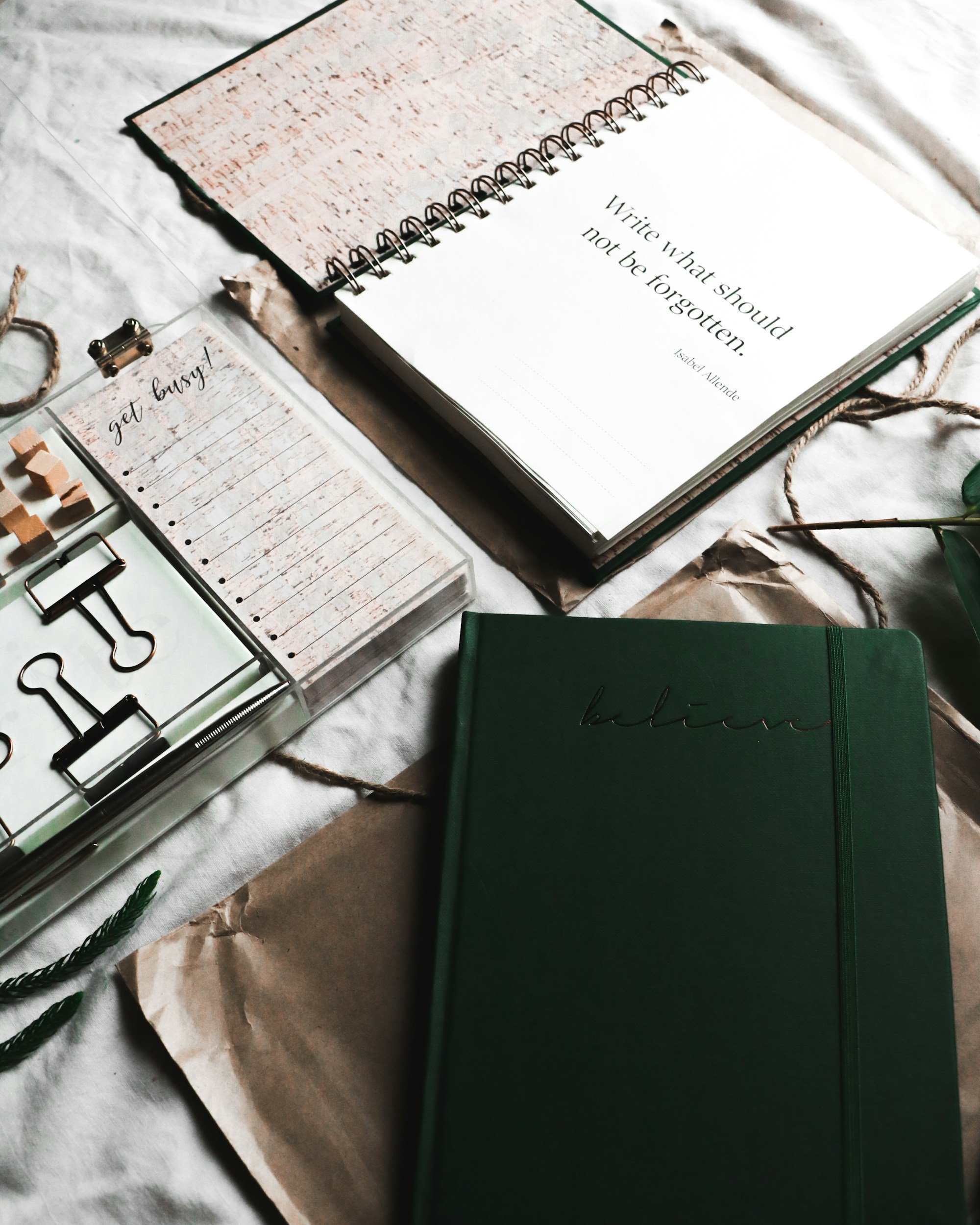 A flat lay of stationery including notebooks, a clear organizer box with paper clips, a notepad labeled 'get busy!', and a paper with the quote 'Write what should not be forgotten' on a white cloth background.