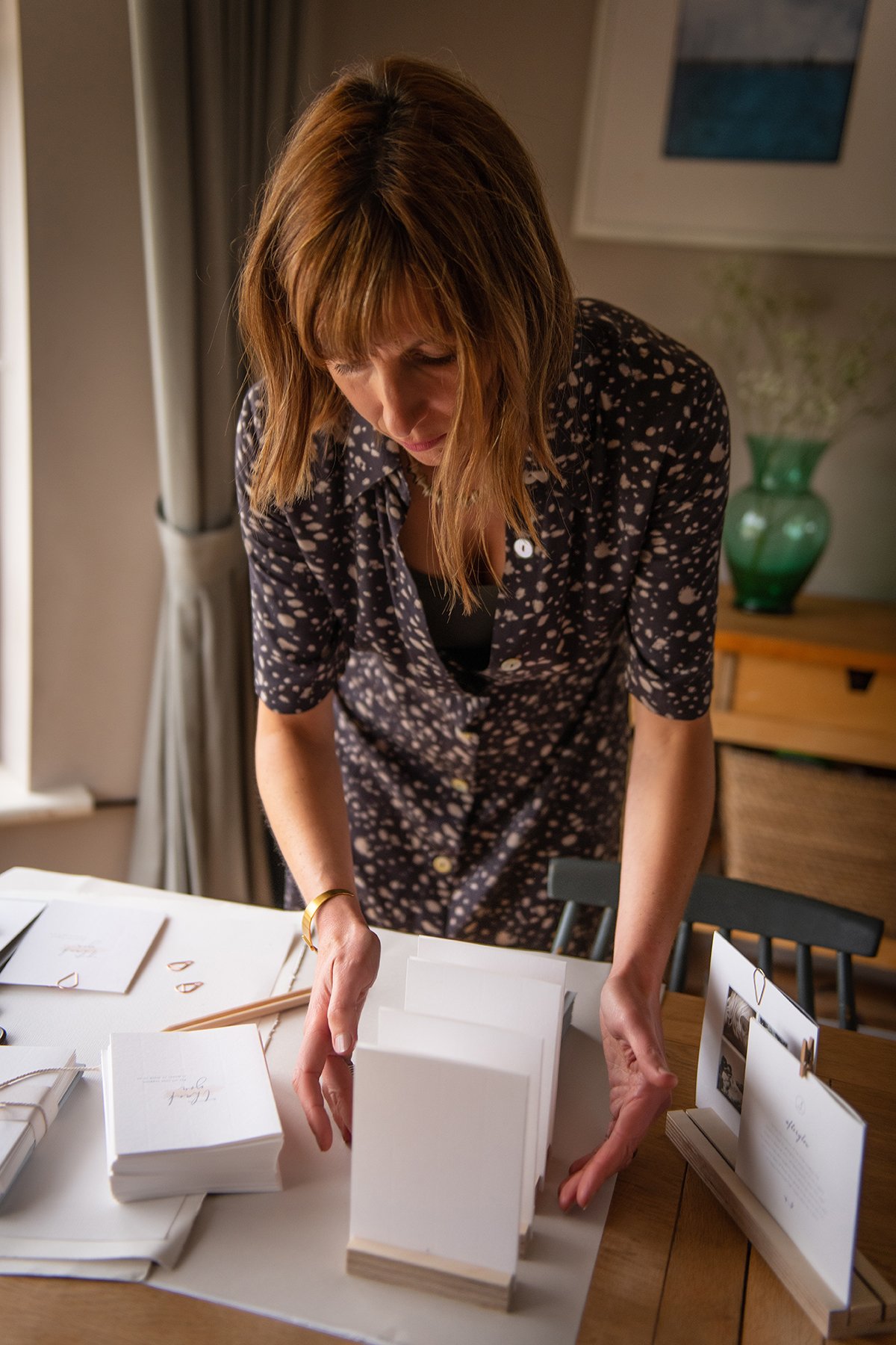 A woman with brown hair, wearing a black and white patterned shirt, is standing at a table arranging paper items.