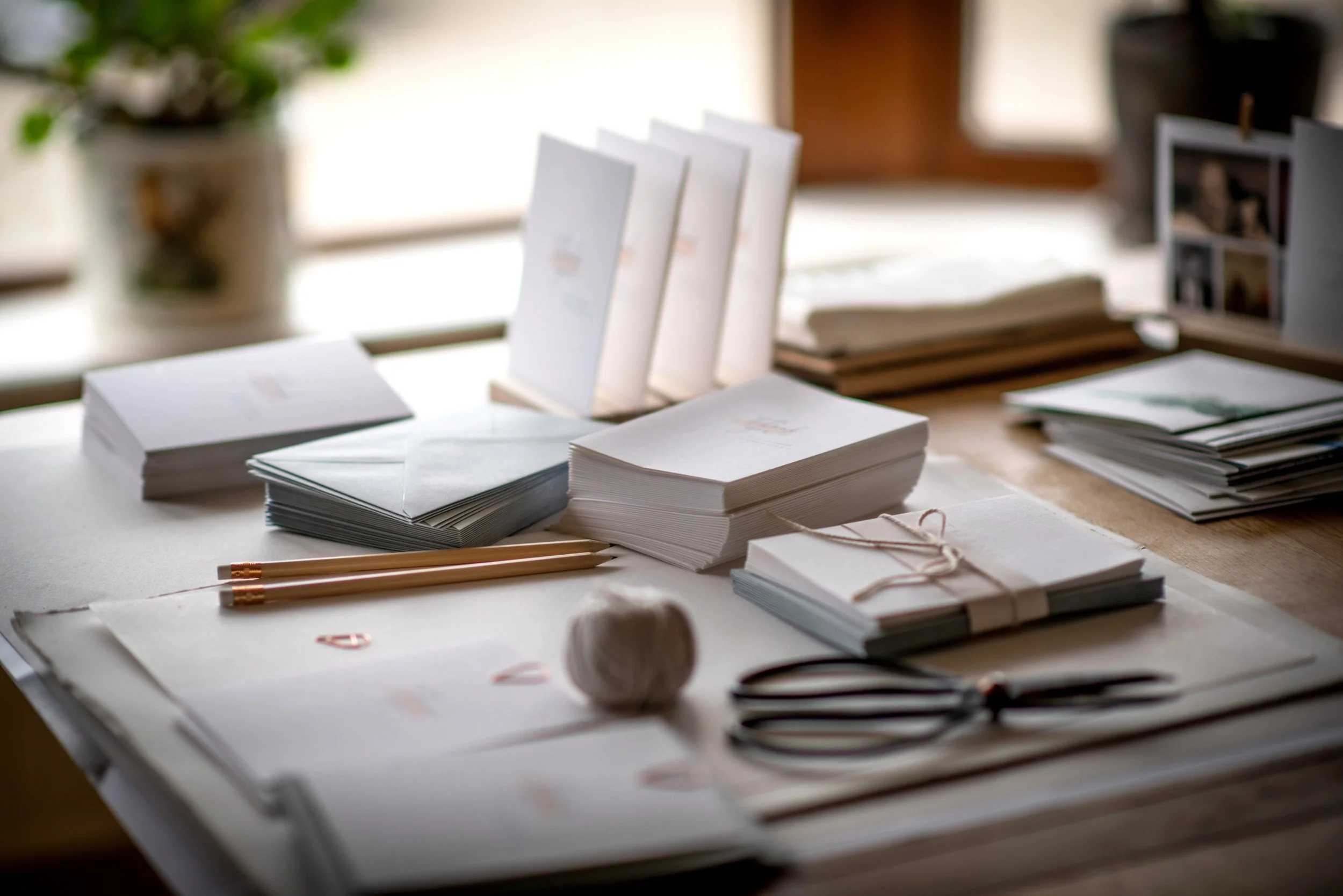 A table with stacks of white paper, envelopes, a ball of twine, scissors, pens, and greeting cards, with a blurred background of a window and a potted plant.