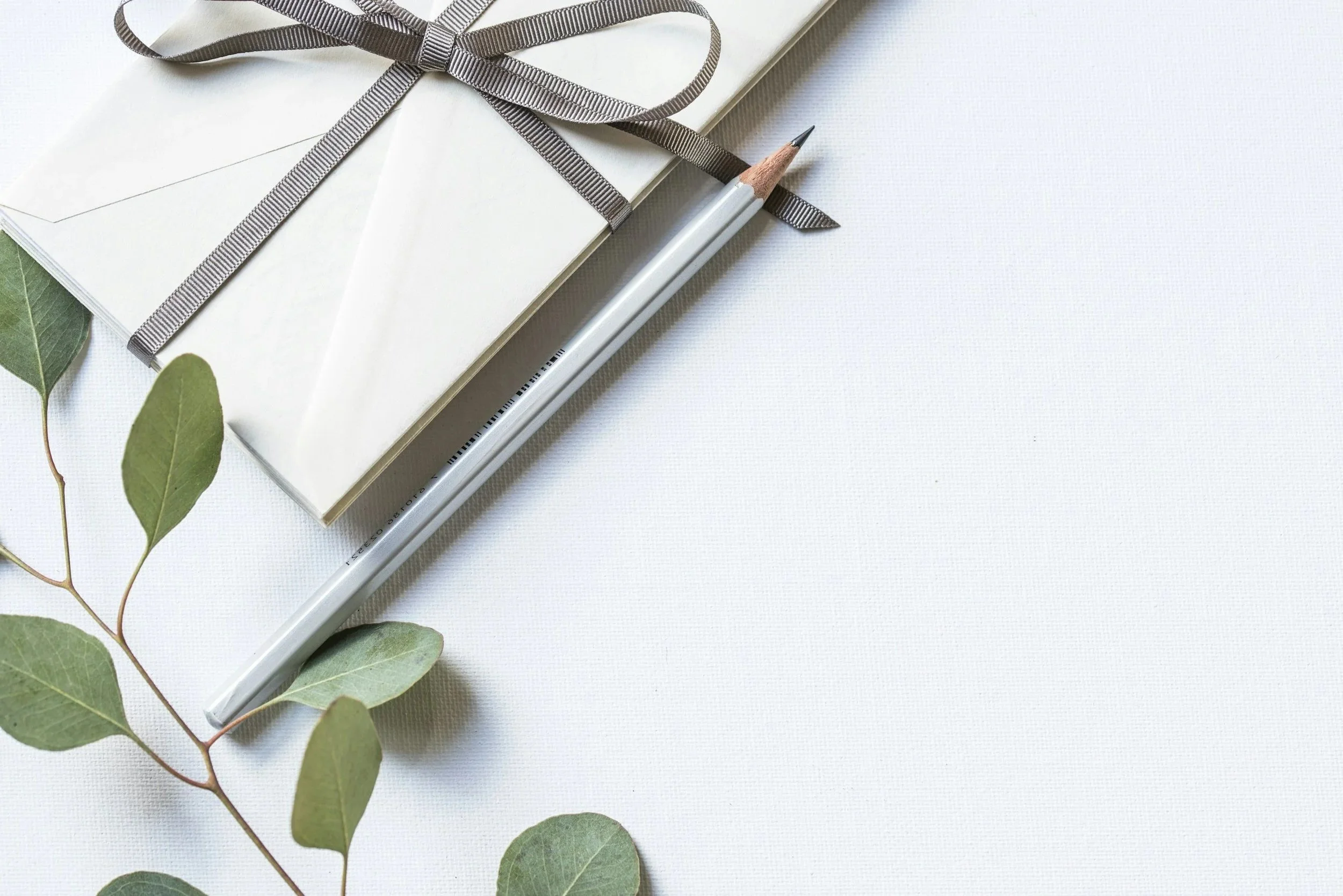 A white wrapped gift with a gray ribbon bow, a white pencil, and some green leaves on a white textured surface.