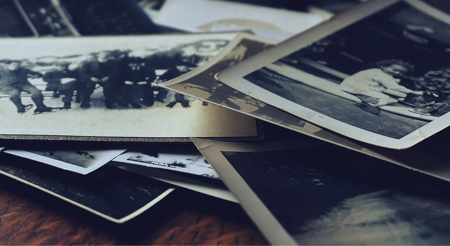 A wooden table covered with old black and white family photographs, carefully laid out while choosing an image for a memorial card.