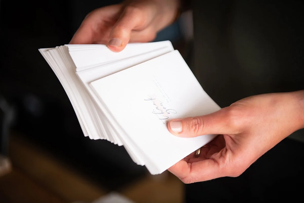 A person holding a stack of white paper cards or booklets, with the top card displaying handwritten text.