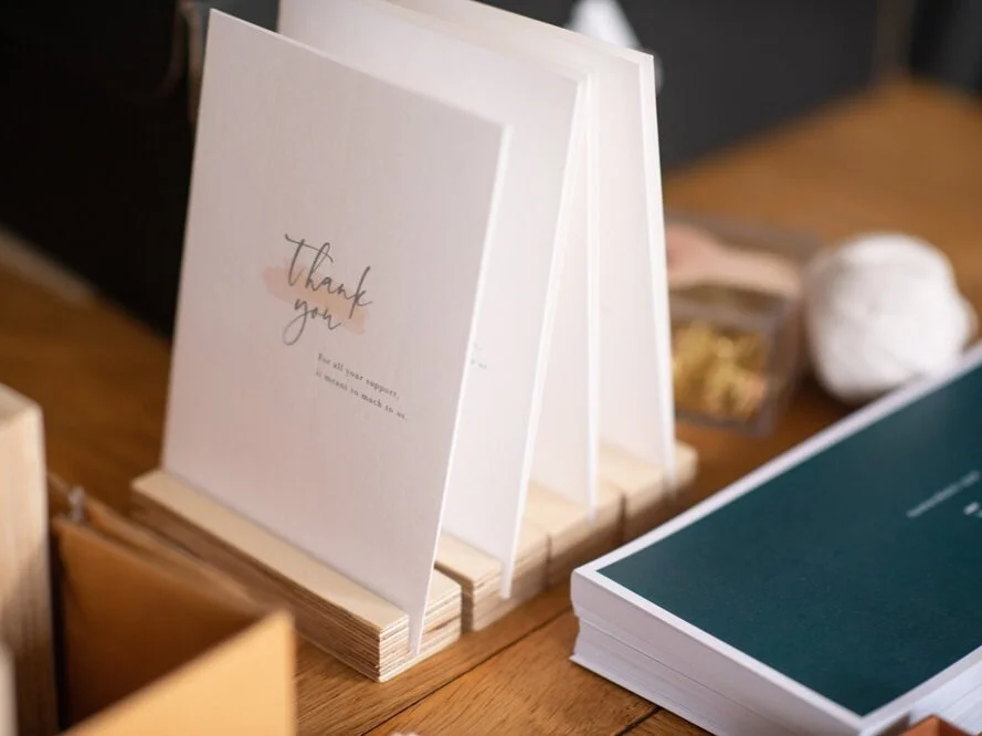 A thank you greeting card displayed vertically on a wooden table, with a faint heart illustration and elegant cursive text that says "Thank you".