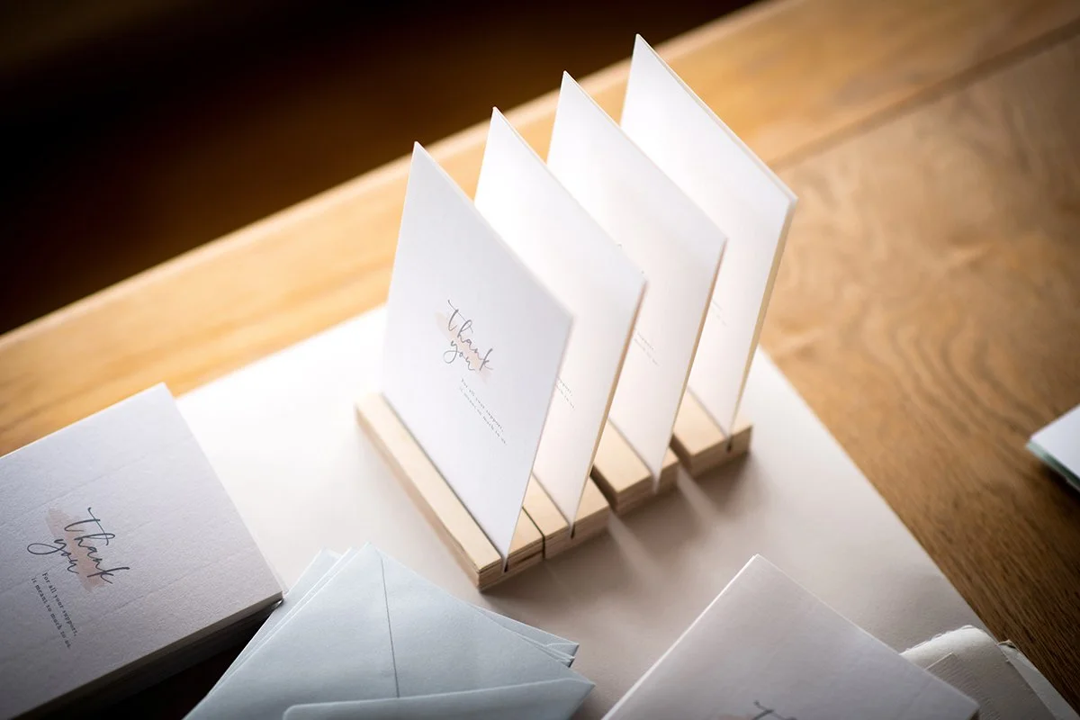 Set of white thank you cards with handwritten style text, displayed on a wooden stand on a wooden table.