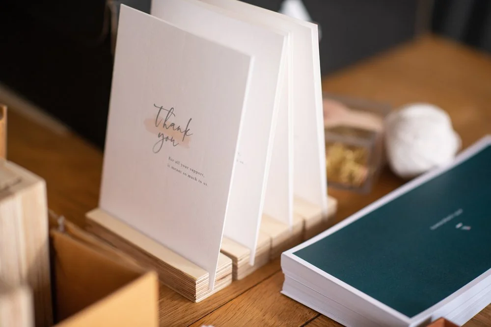 A thank you card standing on a wooden surface next to a large teal-colored booklet and a small container, with a crumpled tissue or paper in the background.