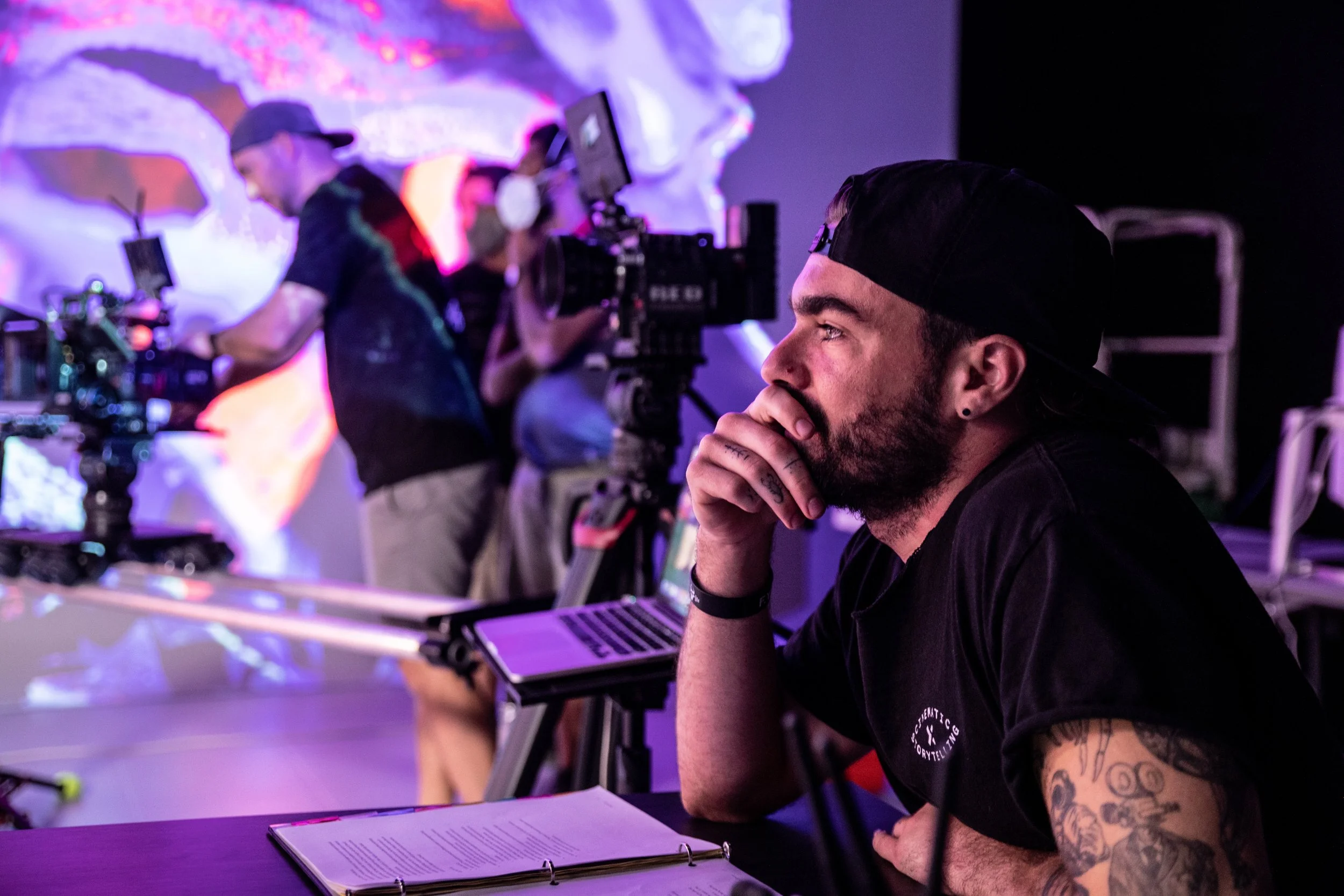 A man with tattoos on his arm, wearing a black shirt and cap, sitting at a table with a folder and laptop, watching a live production or filming with multiple camera operators and crew members working in the background.