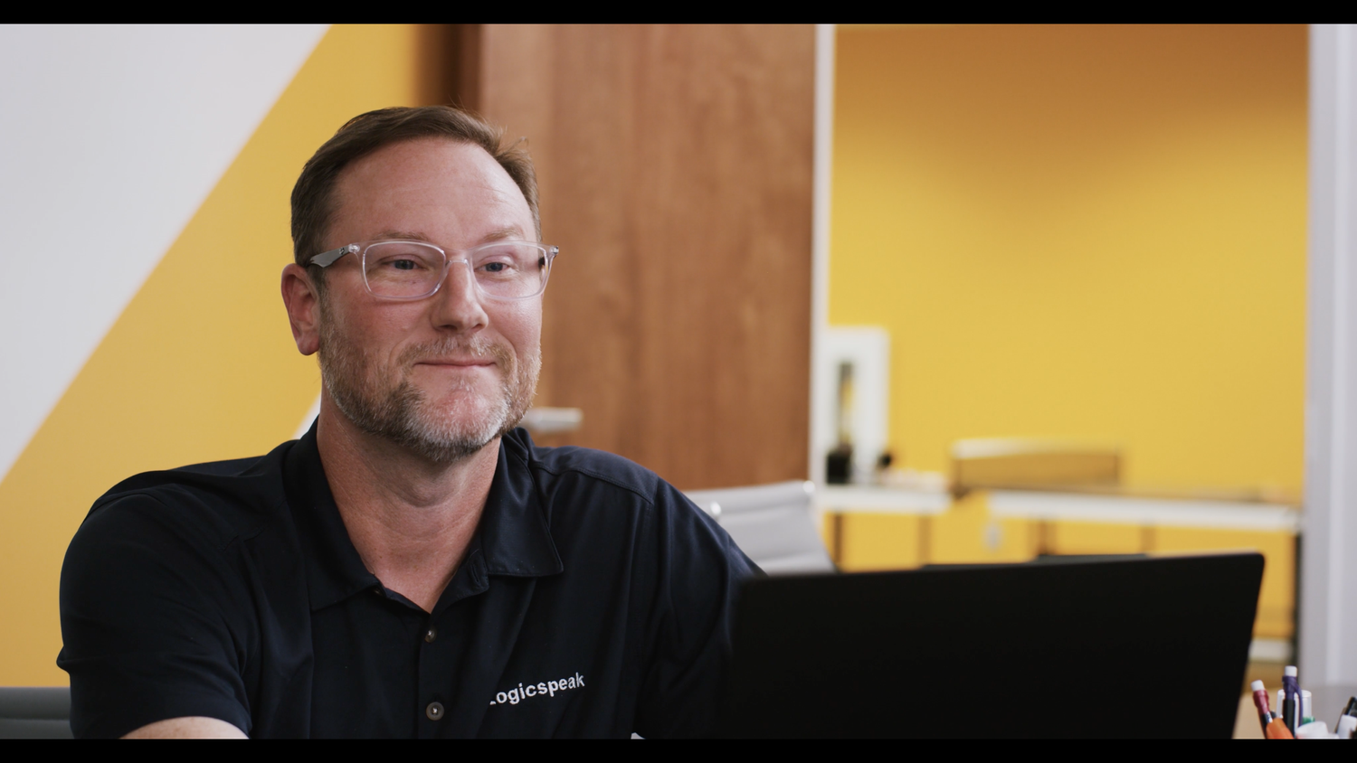 A middle-aged man with glasses and a beard, wearing a black shirt with 'logicspeak' embroidered on it, sitting at a desk in a room with yellow, white, and wood-paneled walls, smiling while looking at a computer screen.