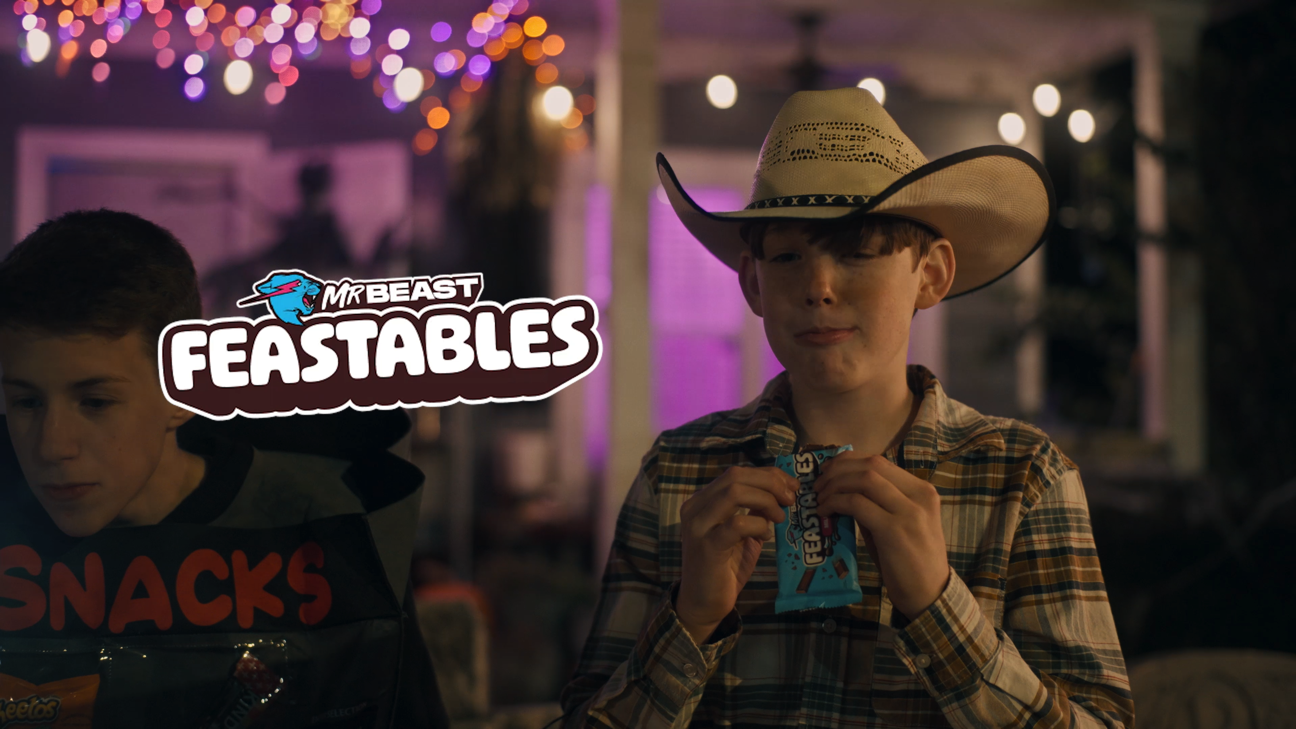 A young boy wearing a cowboy hat and plaid shirt eating a snack at an outdoor gathering during the evening, with blurred decorative lights in the background, and the logo for Mr. Beast's Feastables in the image.