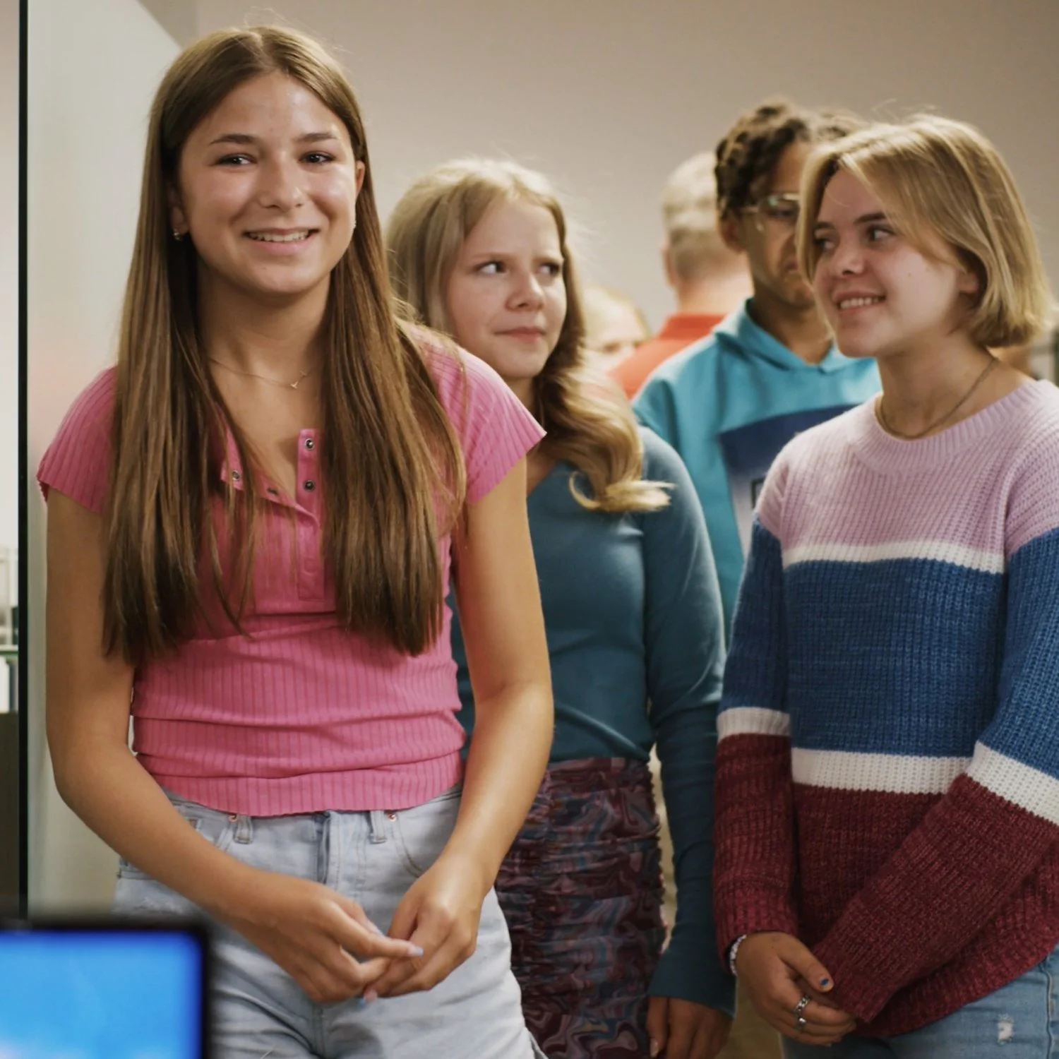 Group of five teenagers standing indoors, smiling and chatting, casual clothing, diverse hairstyles and colors.