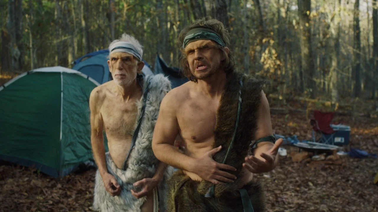 Two shirtless men with headbands and primitive clothing in a forest camp, with tents and camping equipment in the background.