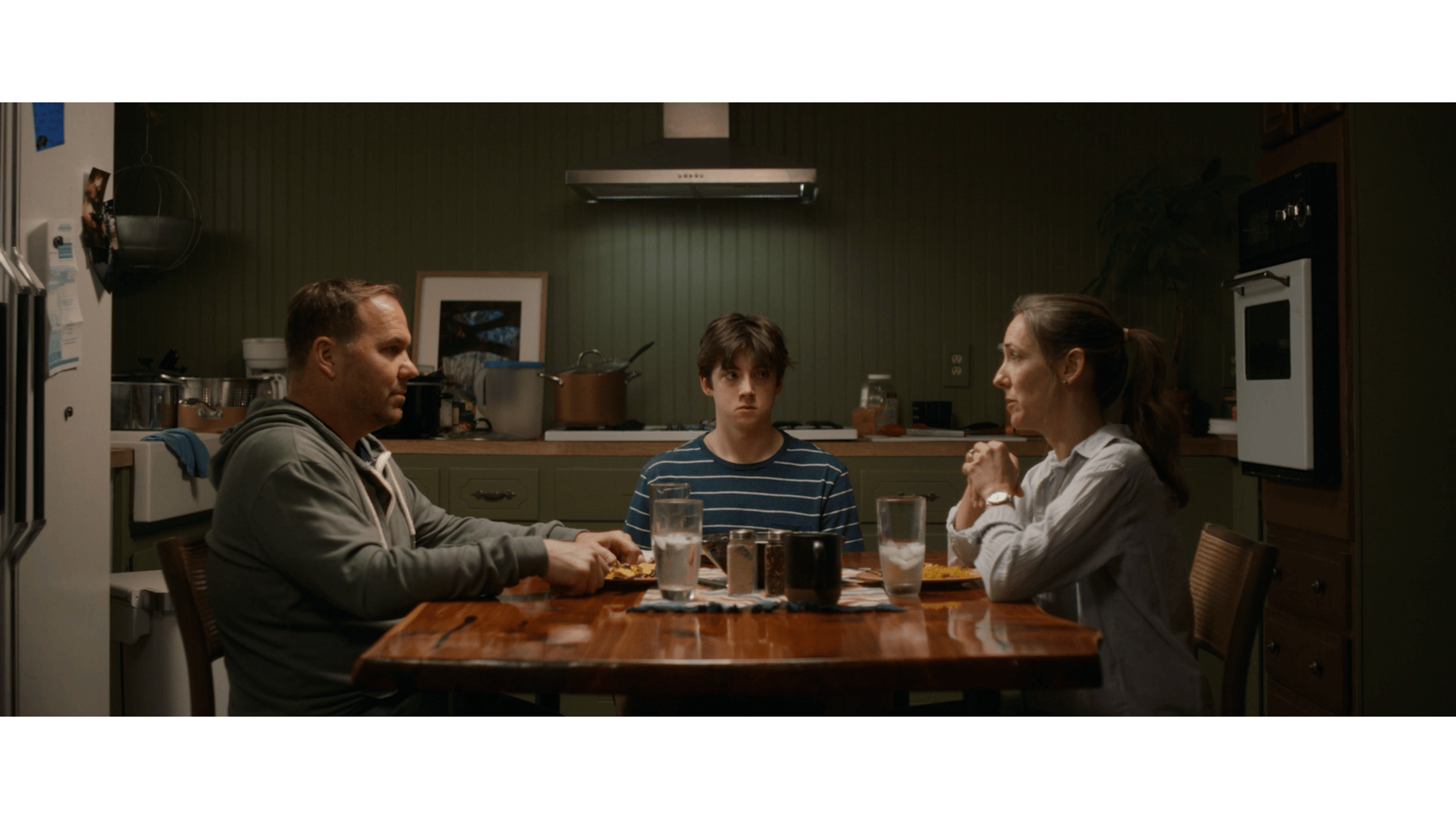A family sits around a dining table in a kitchen. The father on the left, a boy in the middle, and the mother on the right, all engaged in a serious conversation.