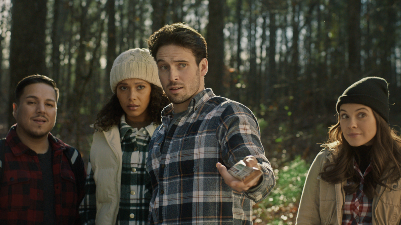 Four young adults standing in a wooded forest, dressed warmly, with serious and curious expressions, with sunlight filtering through trees in the background.