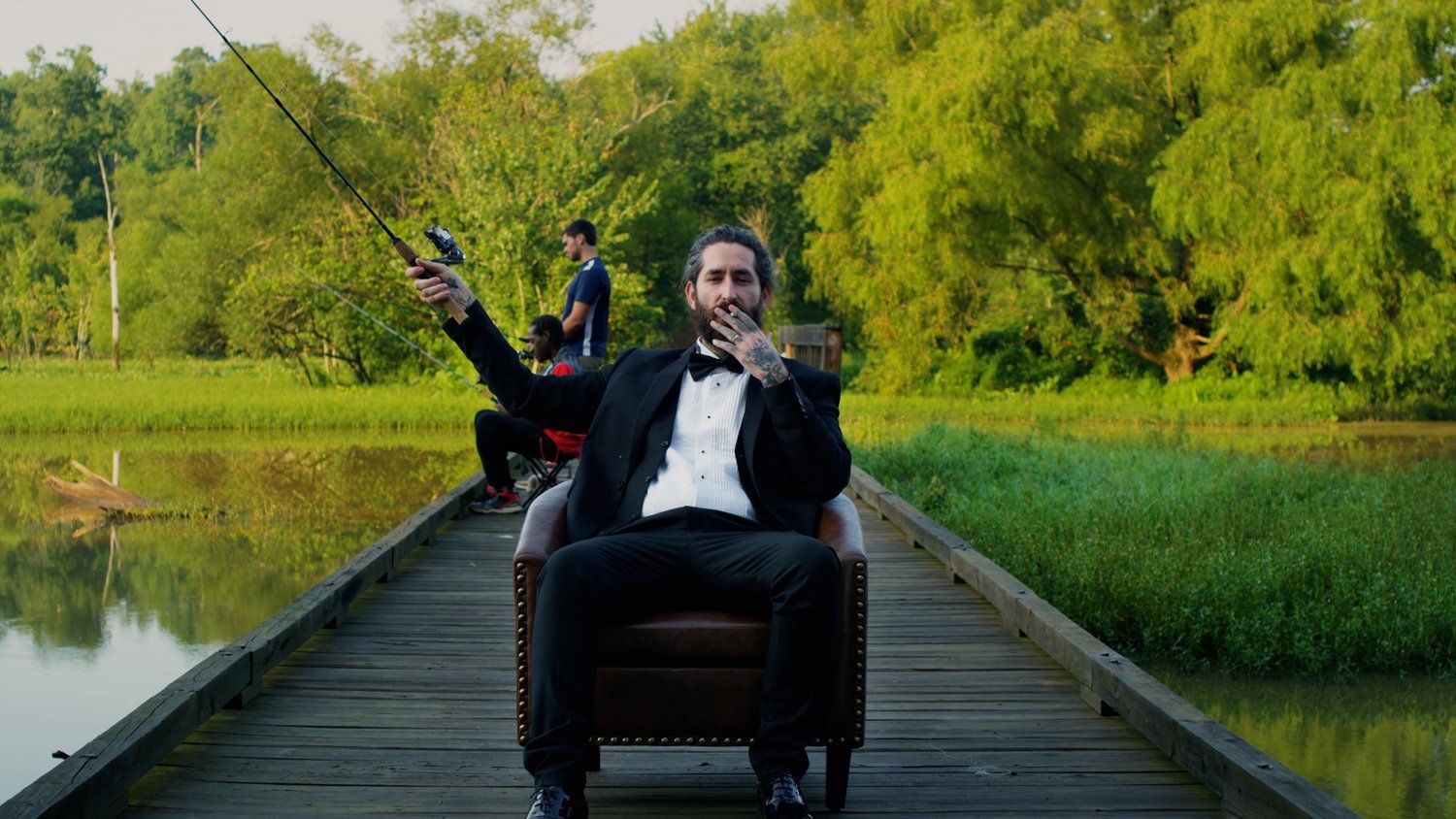 A man in a tuxedo sitting on a chair on a wooden dock while three people fish in the background by a river surrounded by green trees.