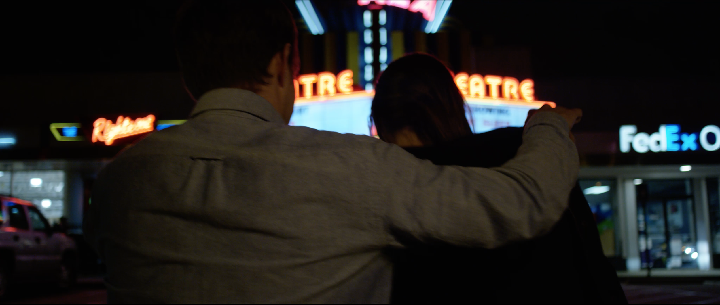 Two people sitting together at night outside a theater with neon signs. One person's arm is around the other, pointing towards the theater.