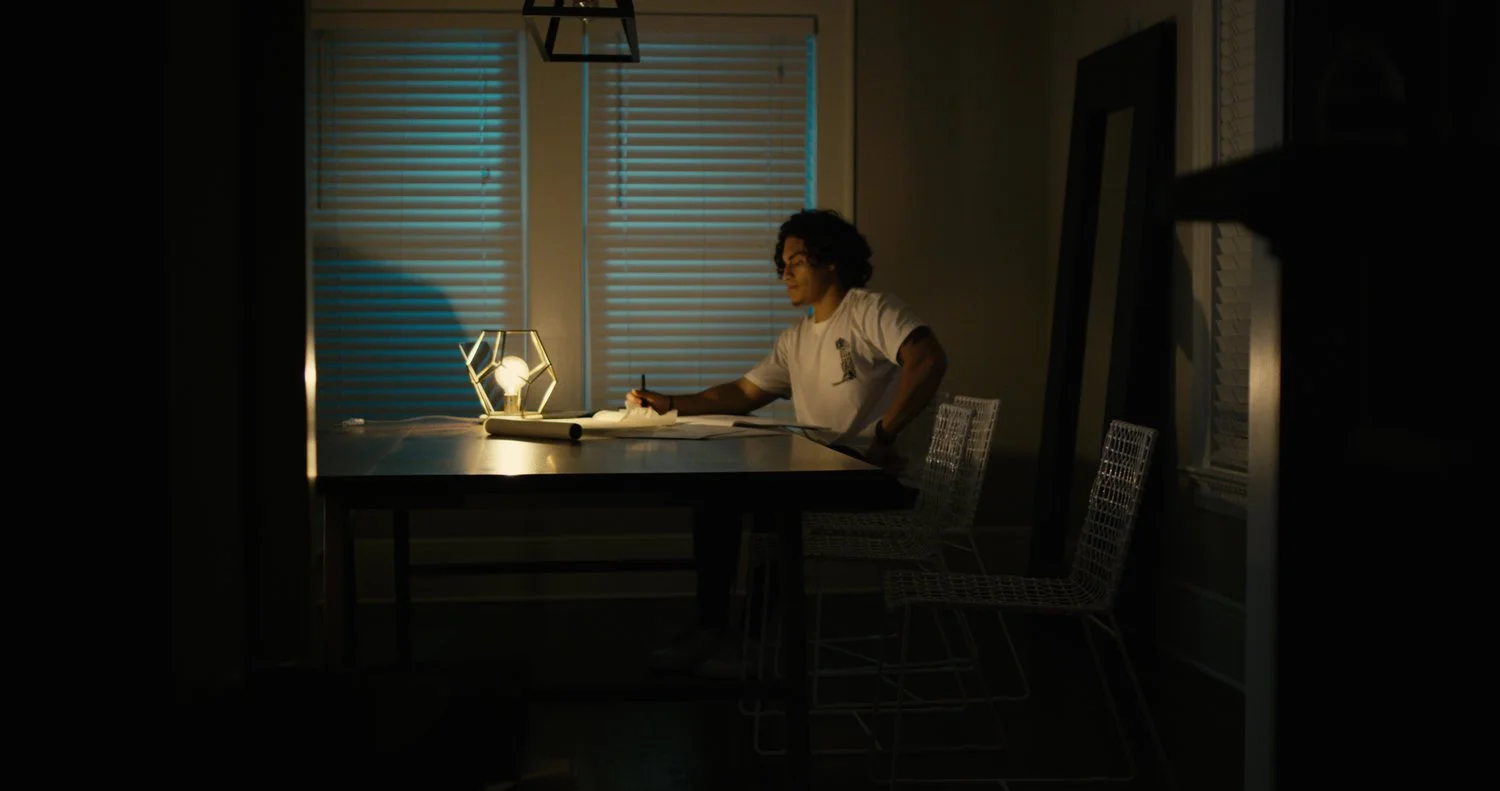 Person sitting at a dining table writing with a glowing decorative lamp, two sets of closed blinds in the background, during evening or night time.