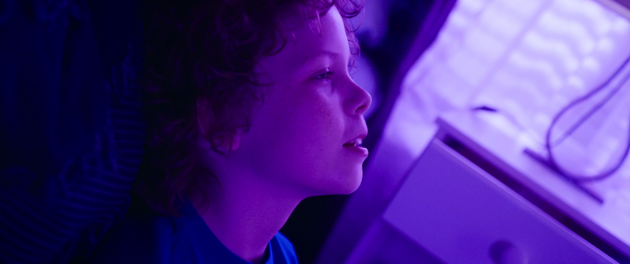 A young boy with curly hair is lying on a pillow, looking at a light projection on the ceiling.