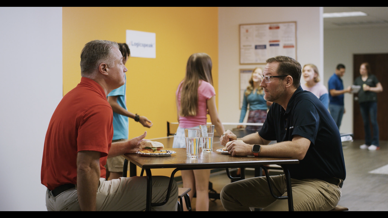 Two men sitting at a table in a cafeteria, facing each other, with glasses of water and a sandwich on the table, engaging in conversation. In the background, several children and teens are talking and walking around in a lively, colorful room.