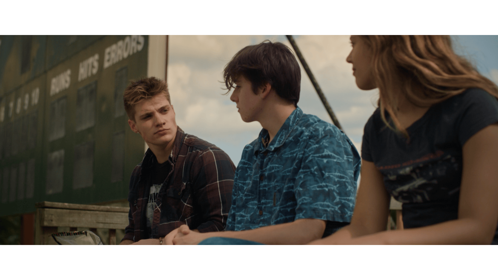 Three teenagers sitting on a wooden bench outdoors, engaged in conversation, with a green building and cloudy sky in the background.
