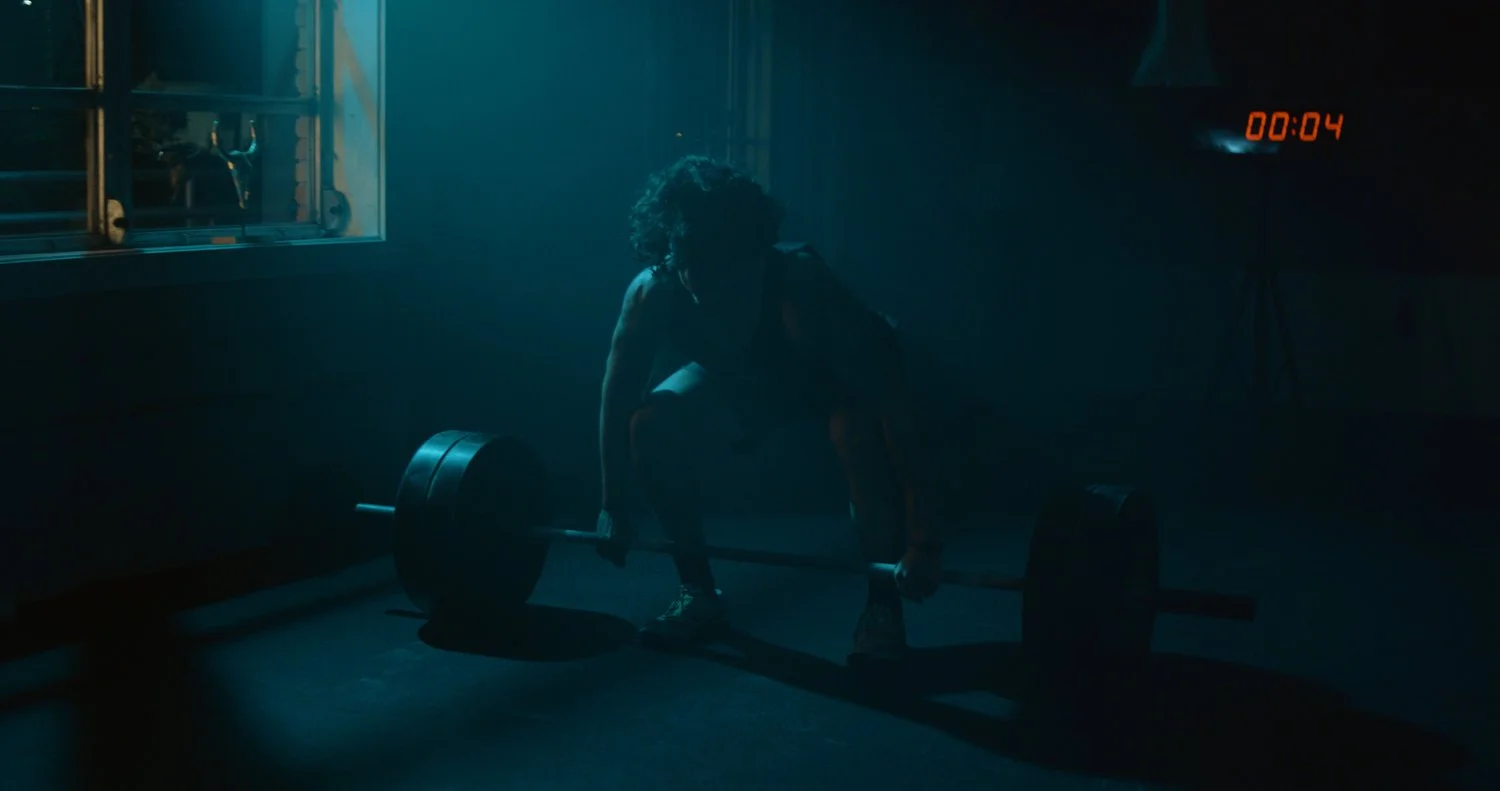 A person with curly hair prepares to lift a barbell with weights in a dimly lit gym, with a timer showing 4 seconds in the background.