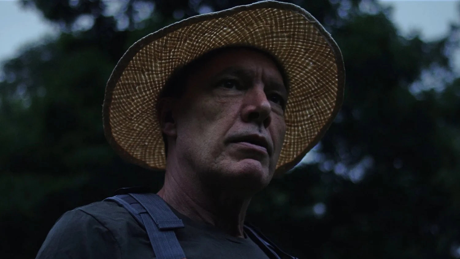 A man wearing a straw hat and dark shirt, looking serious, outdoors in a natural setting with trees in the background.