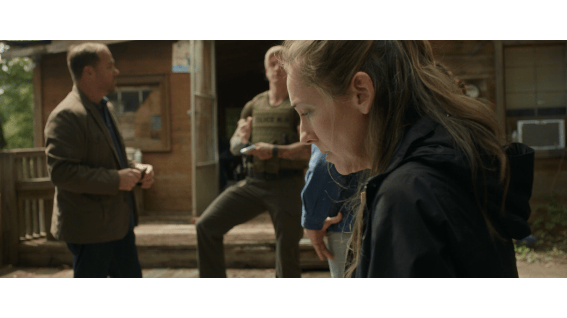 People standing on a porch outside a wooden cabin, with one woman in the foreground looking down, and three men in the background engaged in conversation.