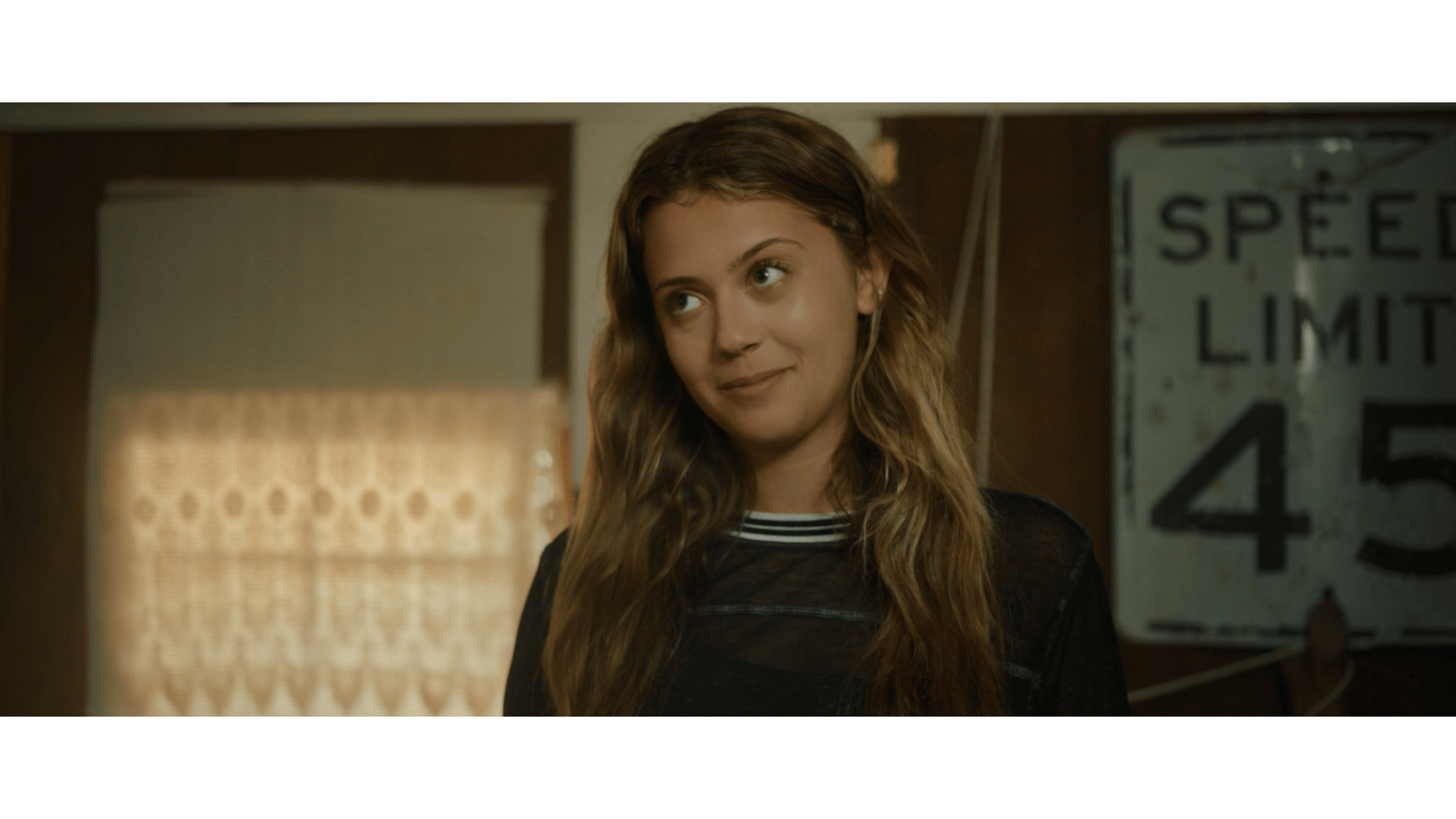 A young woman with wavy brown hair and light skin outside, with a neutral expression, wearing a black translucent top, in front of a wooden wall, a window with lace curtains, and a weathered speed limit sign reading 45.