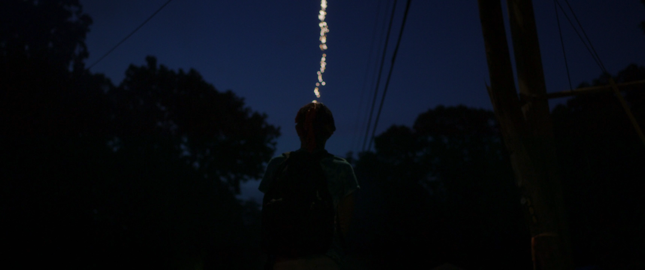 A person standing outdoors at night under a dark sky, looking up at a string of lights hanging above.
