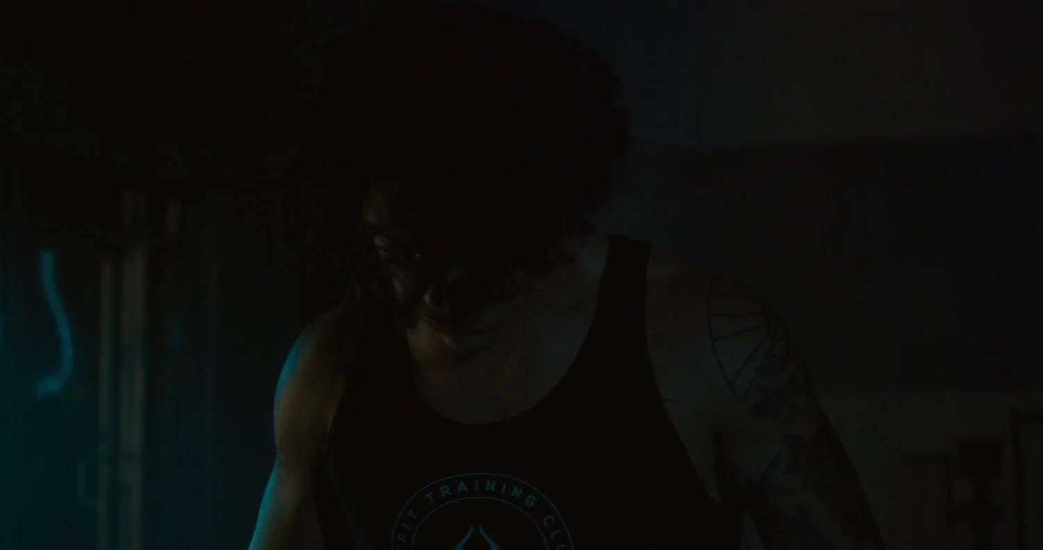 A person with curly hair wearing a black tank top with a logo, in a dimly lit room.