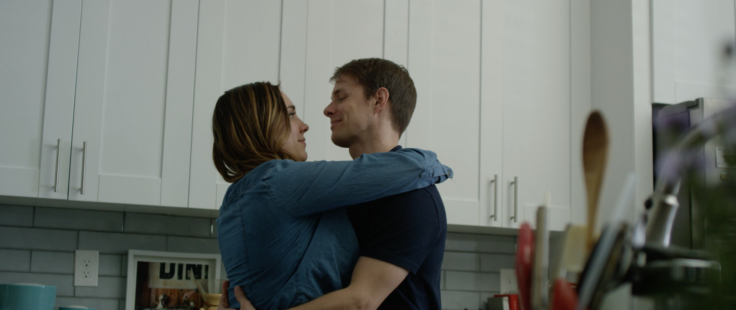 A couple embraces in a kitchen, gazing into each other's eyes with smiles.