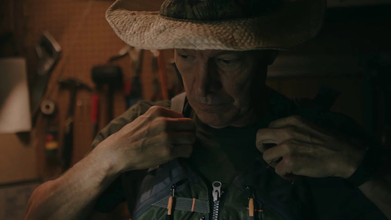 Older man with a straw hat adjusting his backpack in a dimly lit workshop or garage.