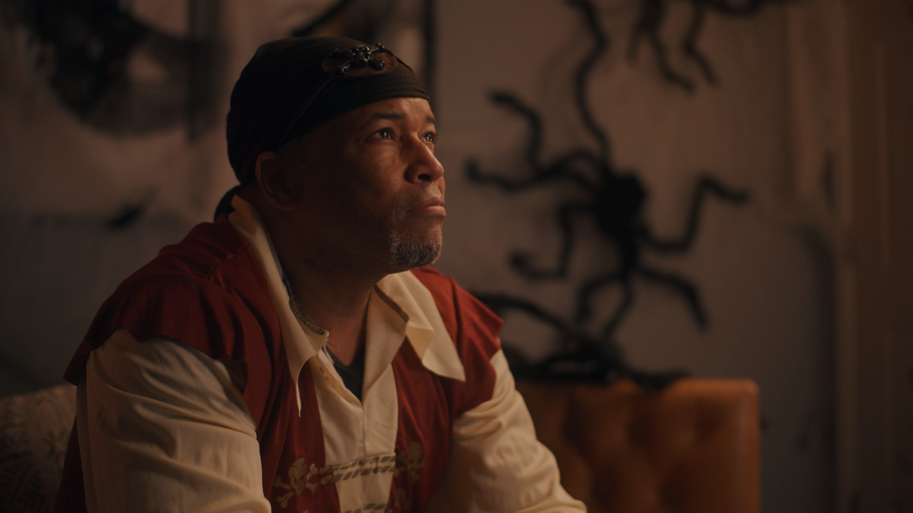 A middle-aged man with dark skin, wearing a black bandana and a tan and red shirt, looking contemplative in a warmly lit room with wall art and a decorative sculpture in the background.