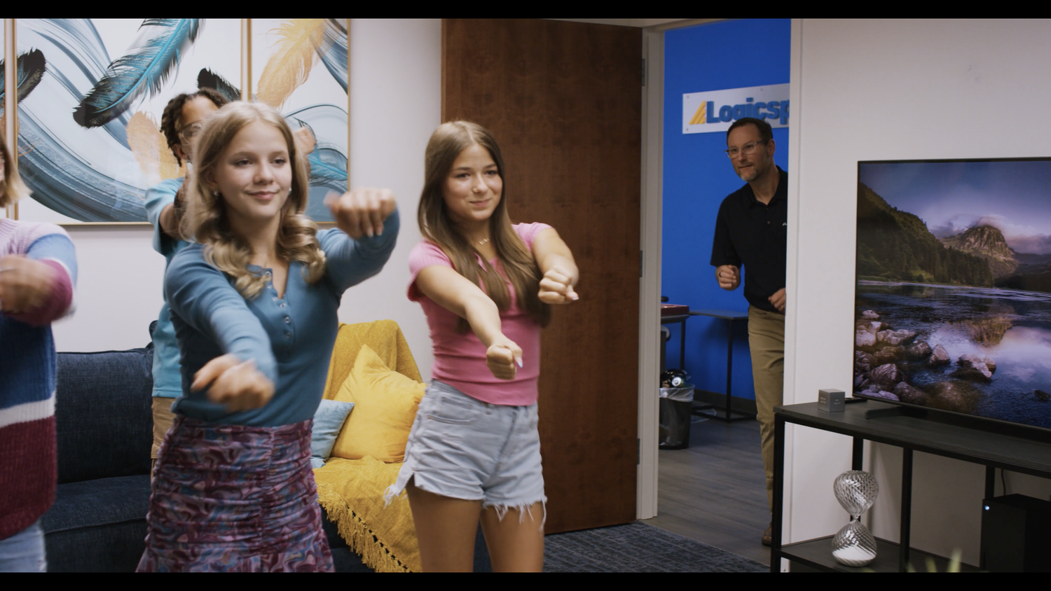 Group of children practicing dance moves in a living room, with an adult man watching from doorway.